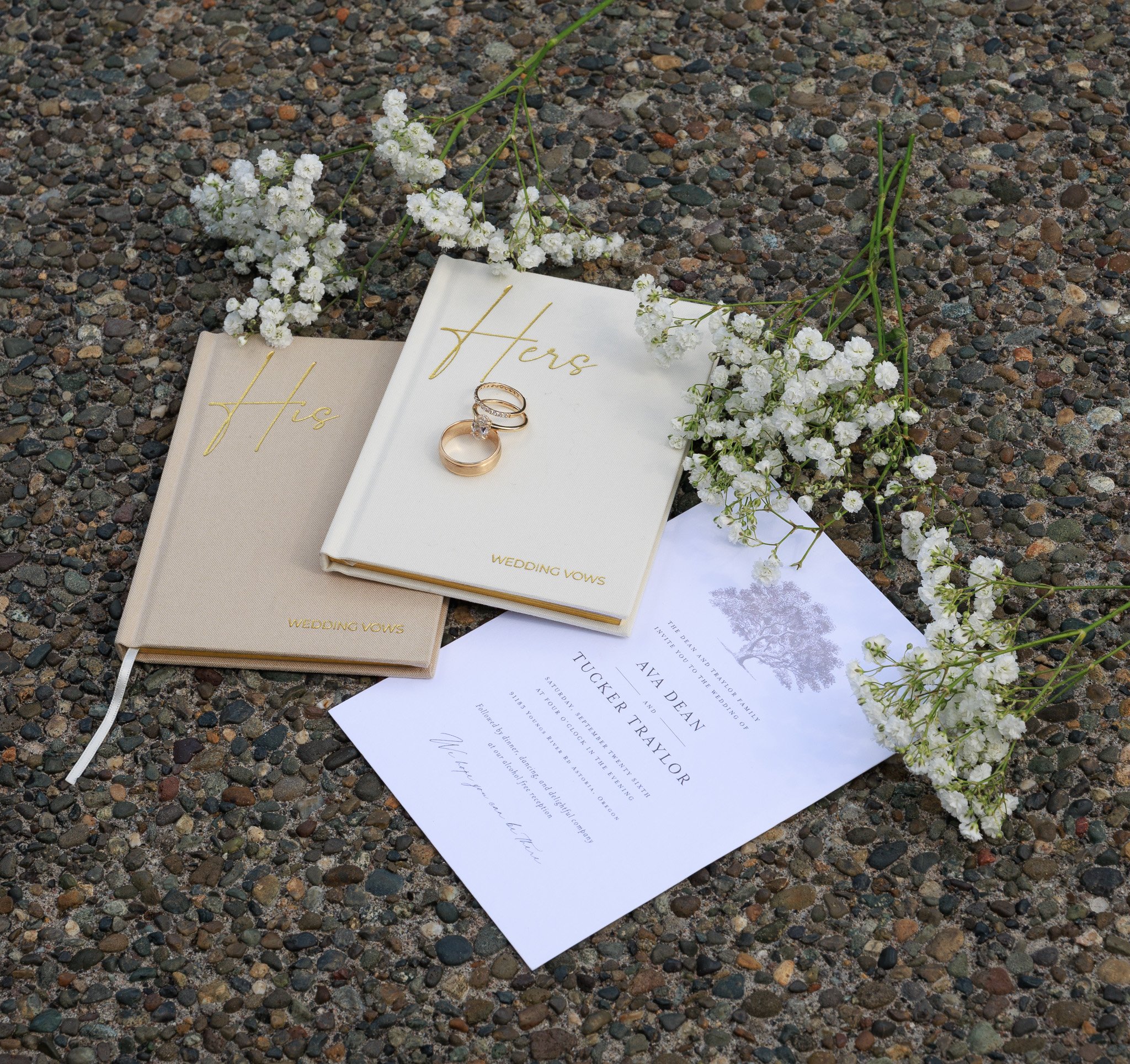 A wedding invitation and rings are placed on a gravel surface surrounded by white floral arrangements. The invitation has gold lettering and the rings include a wedding band and an engagement ring.