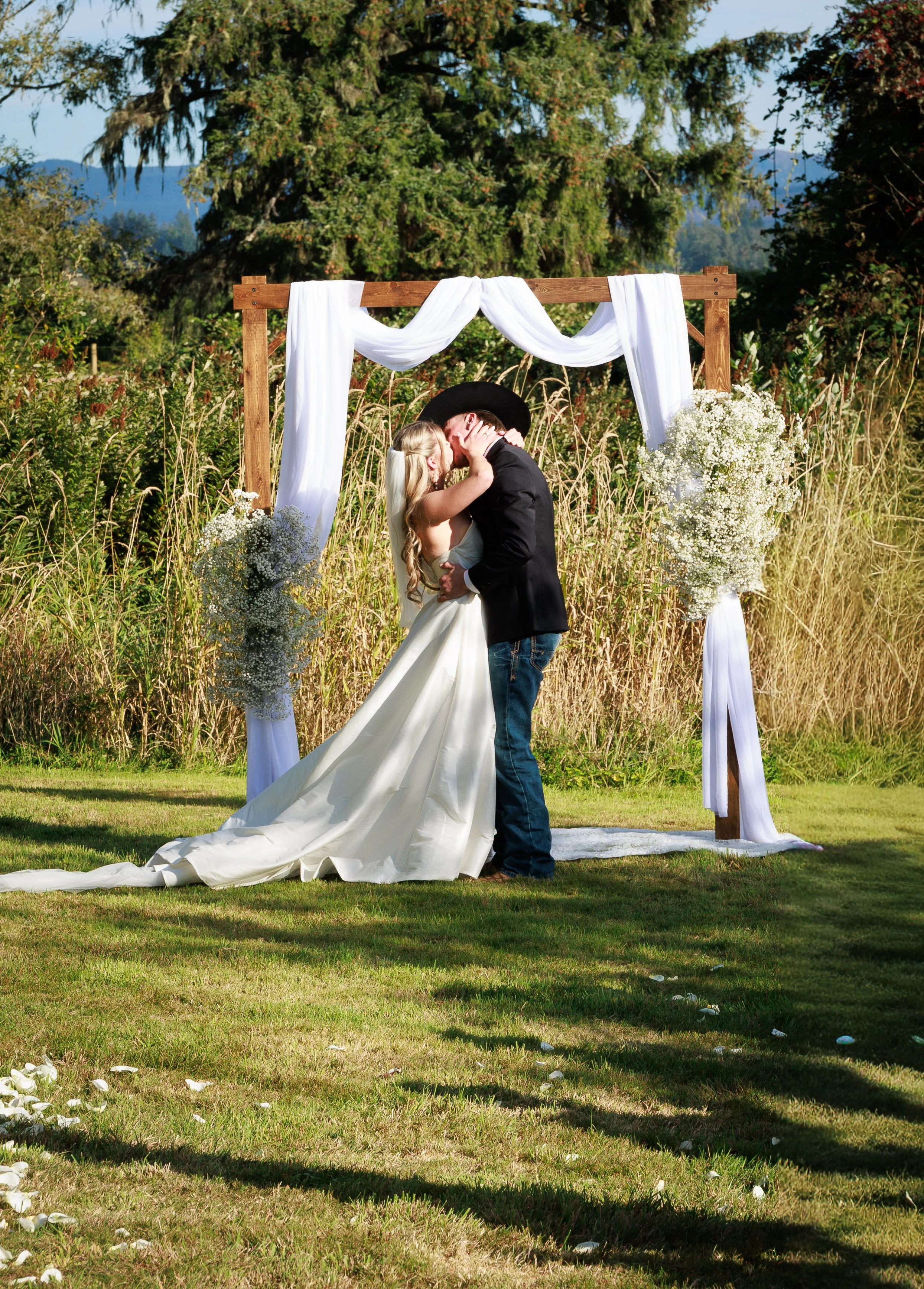 A bride and groom kiss under an outdoor wedding arch decorated with white fabric and flowers in a natural setting with trees and grass.