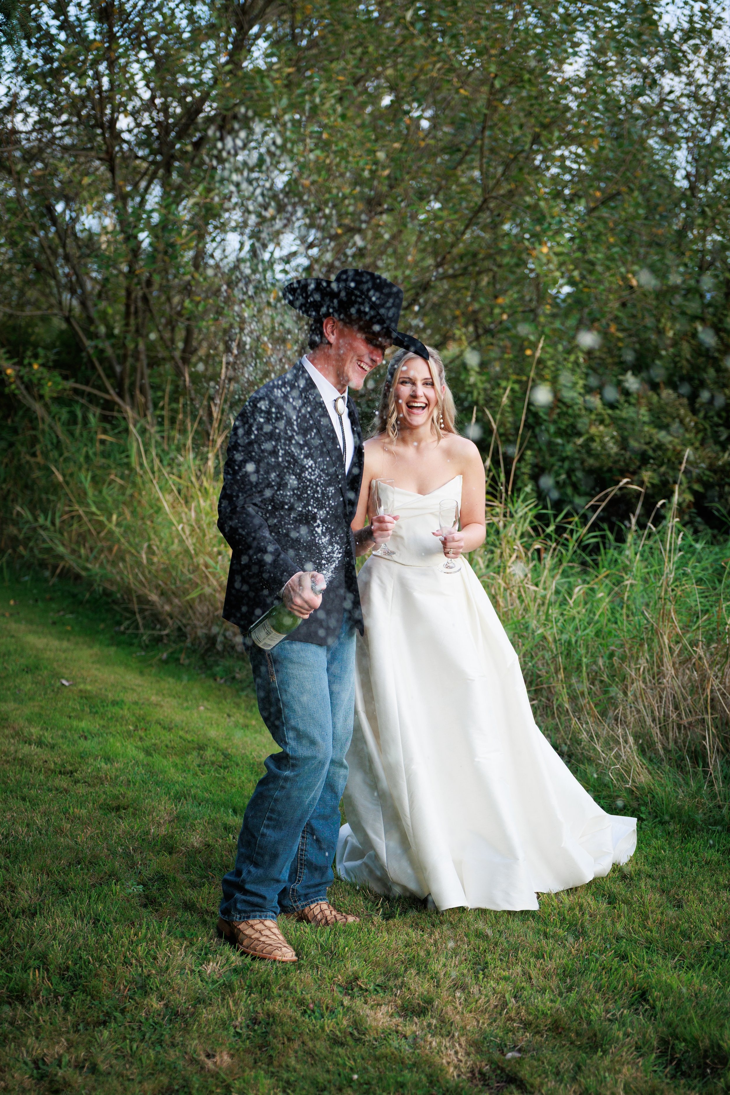 A man and woman celebrating outdoors. The man is wearing a black cowboy hat, black suit jacket, and blue jeans, holding a champagne bottle with foam. The woman is in a white strapless wedding gown, holding a champagne glass, both are smiling and laug