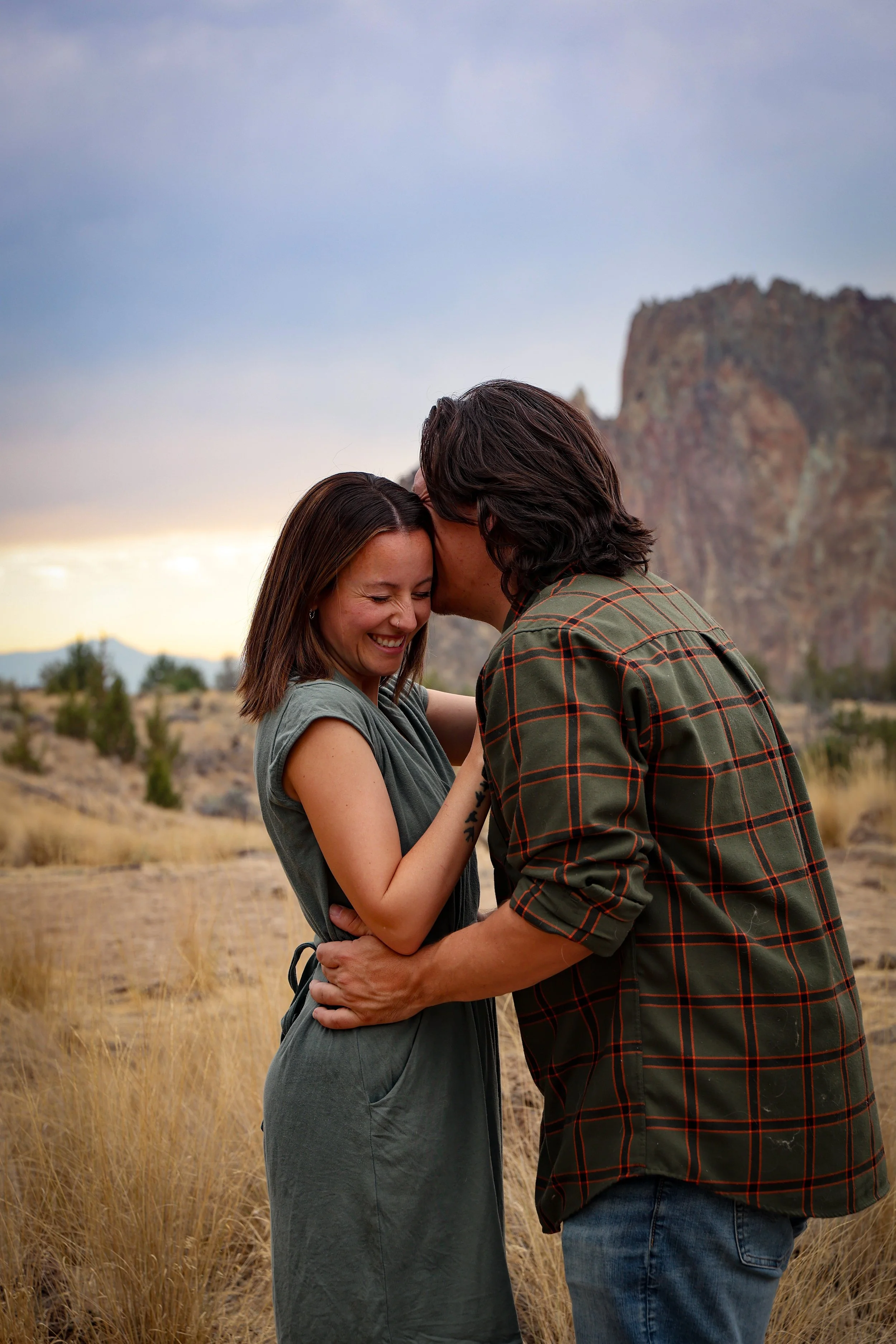 A young woman and a young man are in a field with tall, dry grass, embracing and smiling with their foreheads touching against a rocky mountain and cloudy sky in the background.