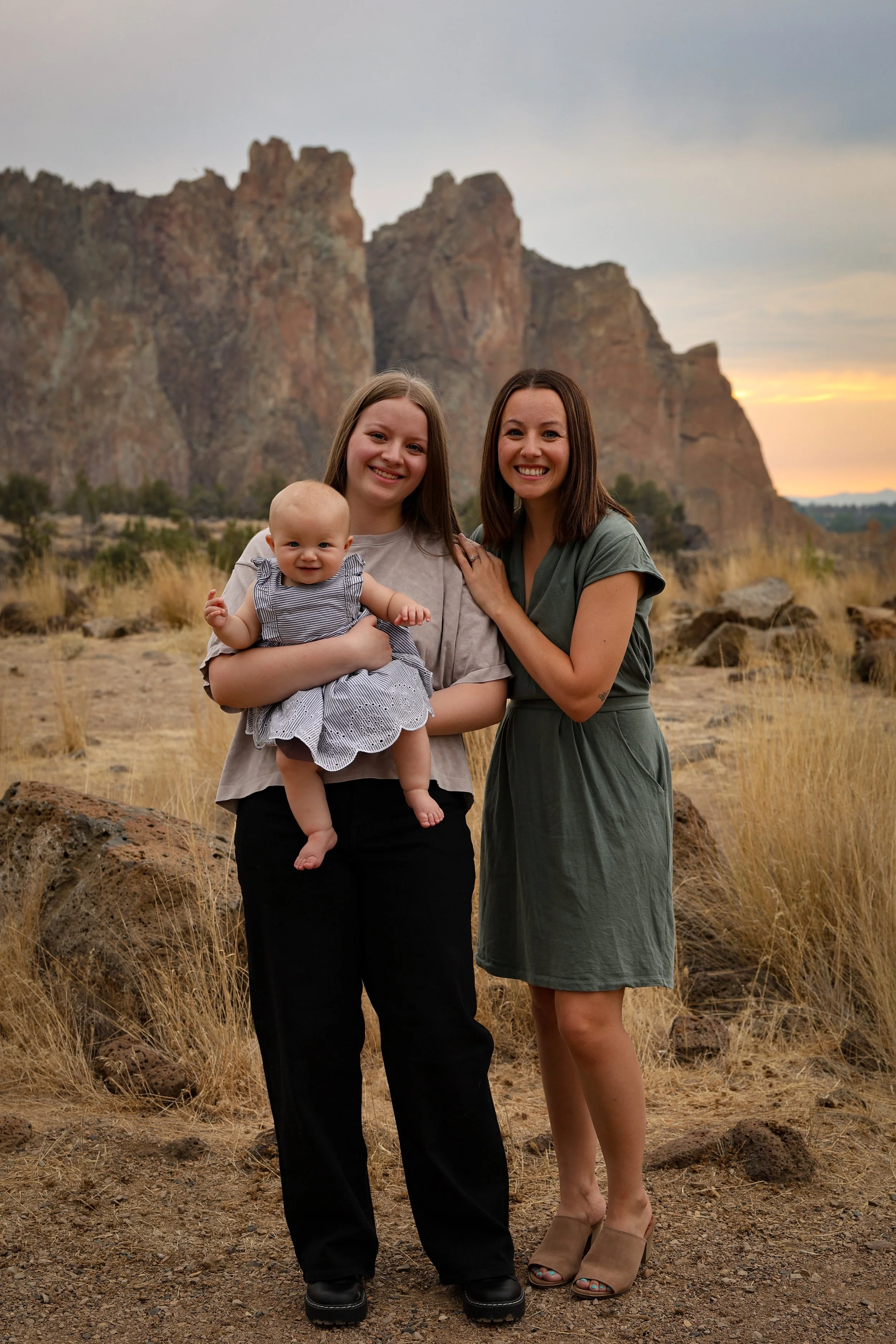 Two women and a baby standing outdoors in front of tall rocky cliffs at sunset, with dry grass and rocks on the ground.