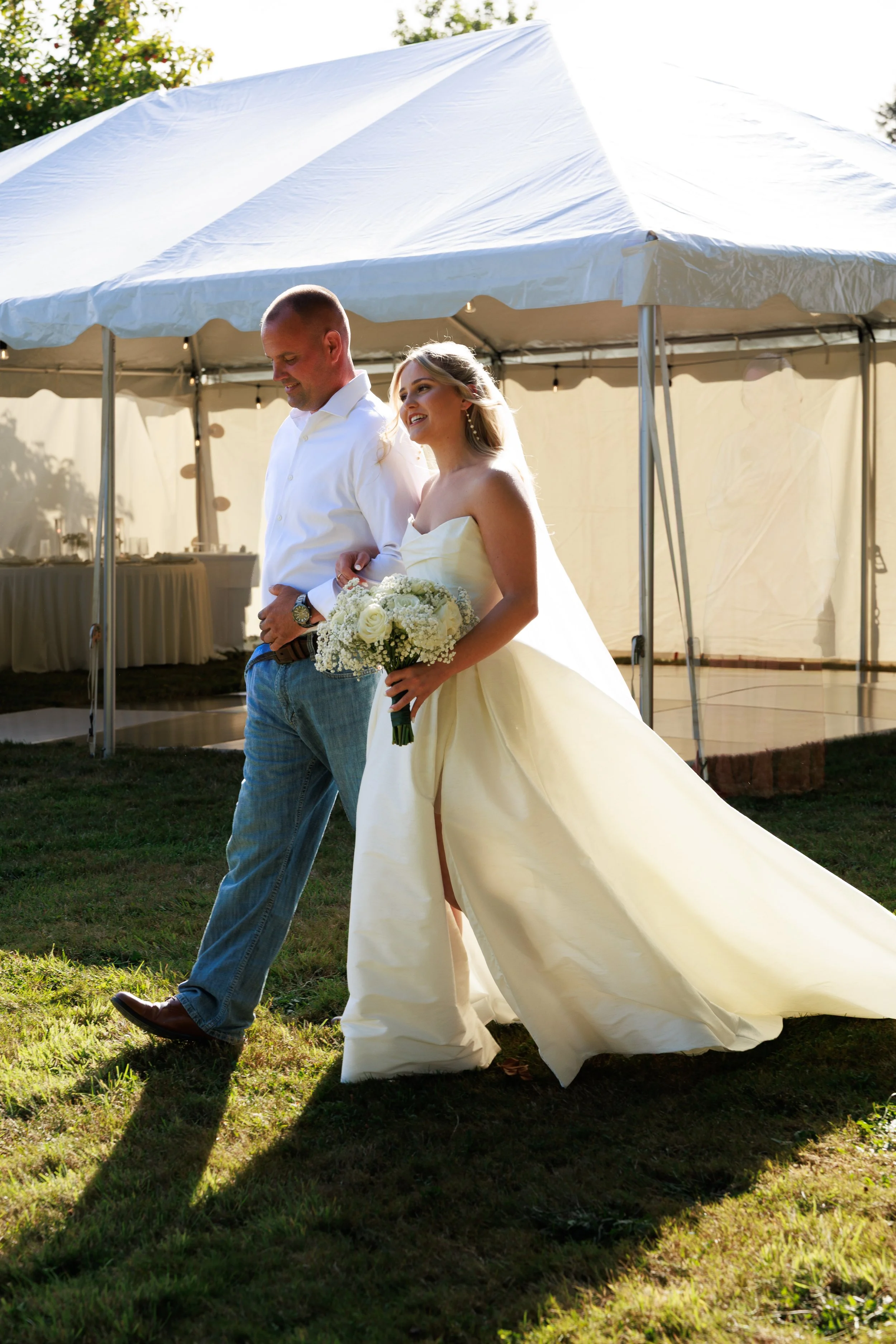 A bride and groom walking together outdoors at a wedding reception, with the bride holding a bouquet of white roses and the groom wearing a white shirt and jeans.