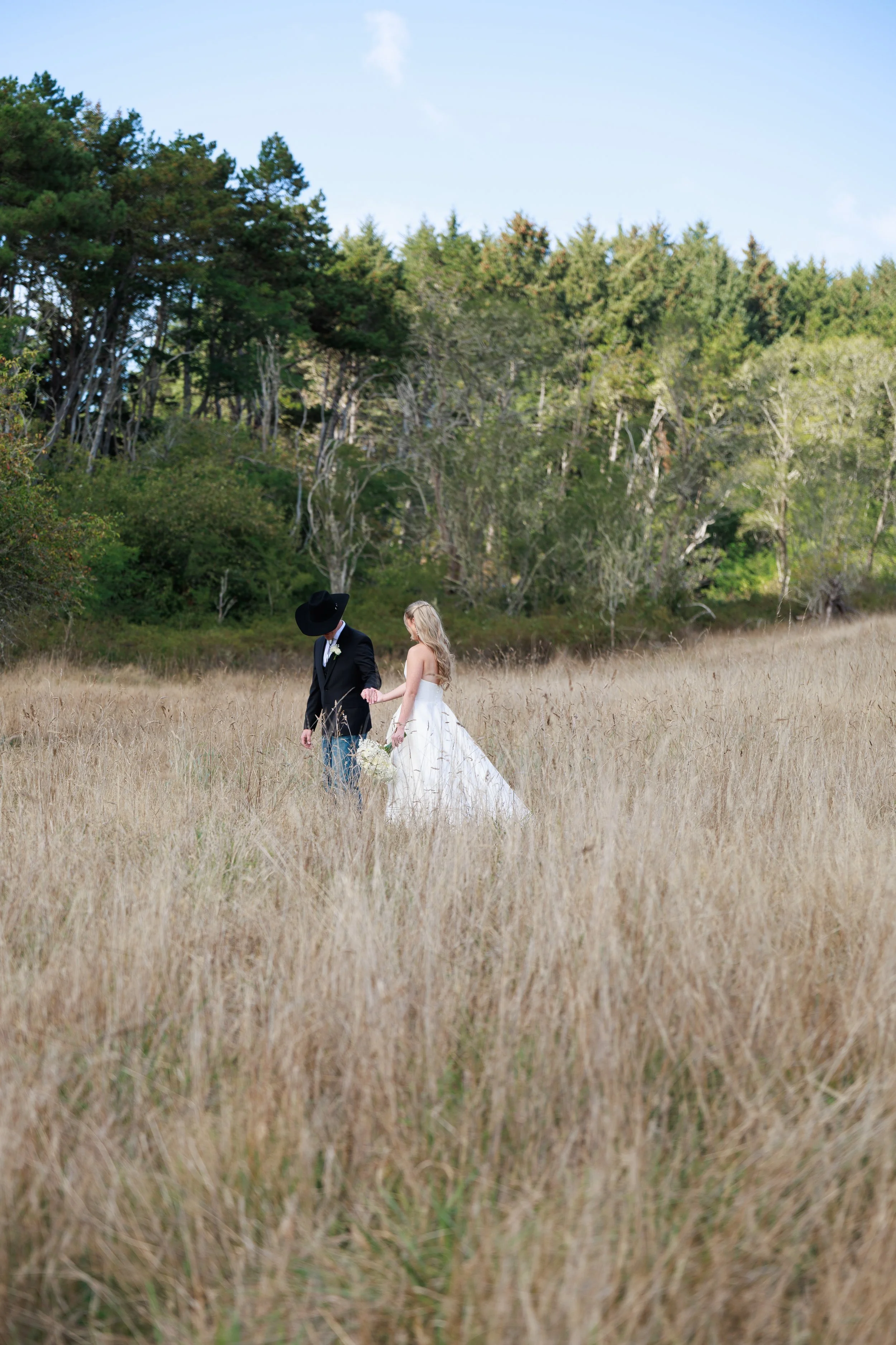 A couple dressed in wedding attire holding hands and walking through a field of tall grass near a forested area.