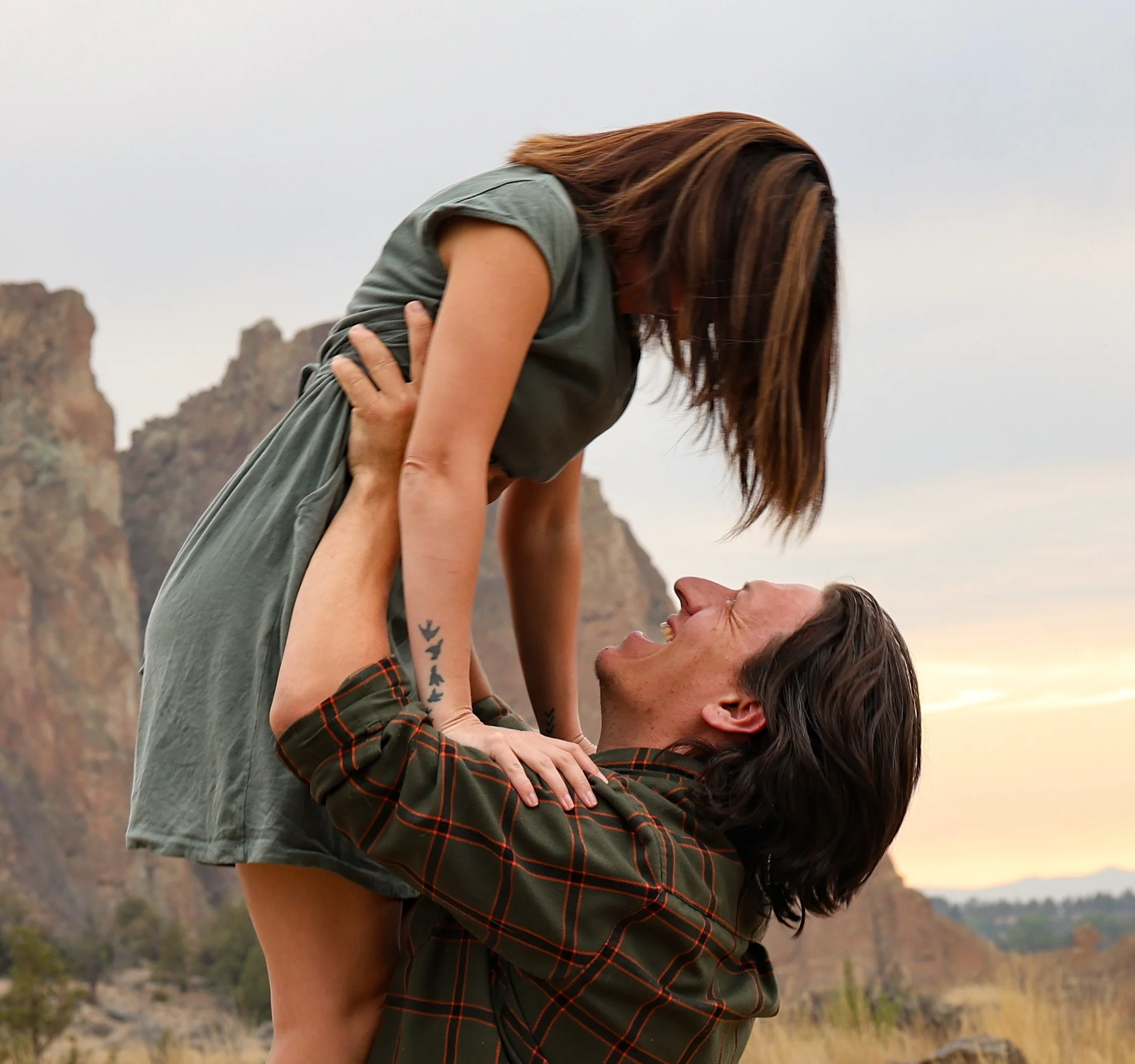 A woman with long, dark hair leaning back, supported by a man lifting her, with mountainous terrain and a cloudy sky in the background.