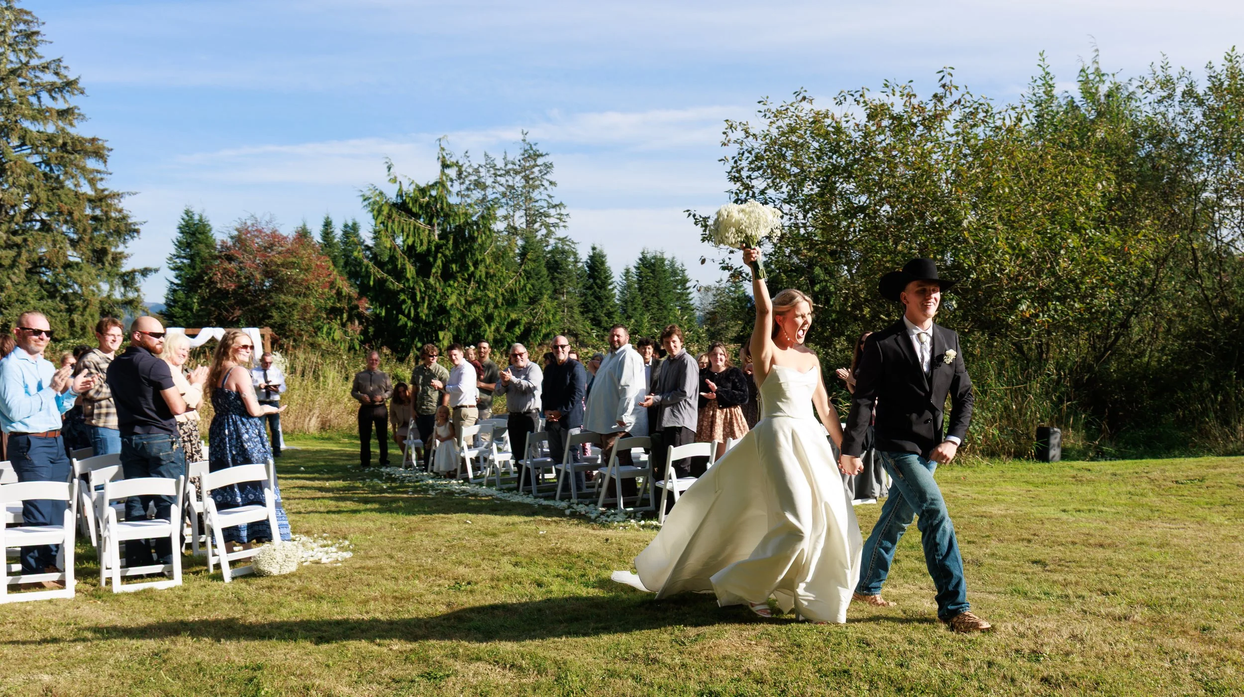 A newlywed couple, the bride in a white dress holding a bouquet, and the groom in a black hat and suit, walking hand-in-hand outdoors with guests watching.