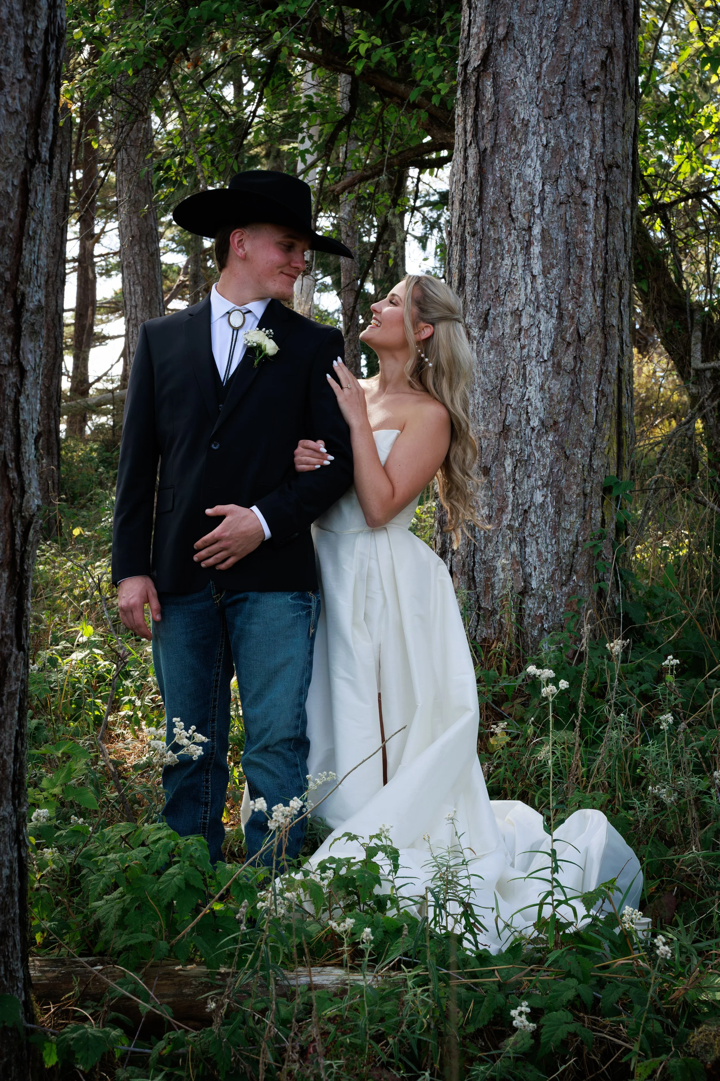A couple dressed in wedding attire standing in a wooded area, the bride in a strapless white gown and the groom in a black suit with a cowboy hat, looking at each other affectionately.