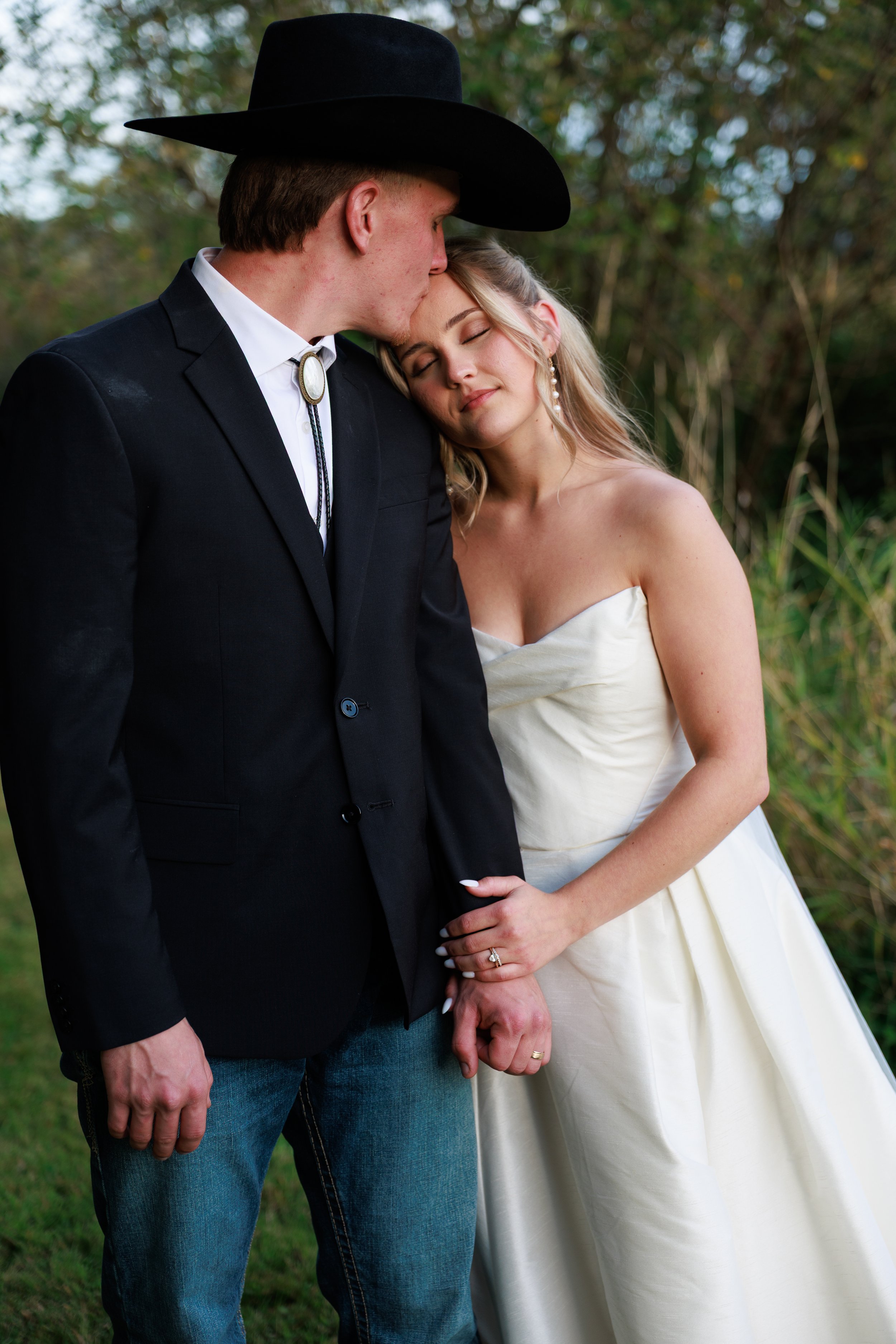 An engaged couple standing outdoors. The man is wearing a black cowboy hat, a black blazer, a white shirt, and a bolo tie. The woman is in a strapless white wedding dress, with her eyes closed and a content expression. They are holding hands, and the