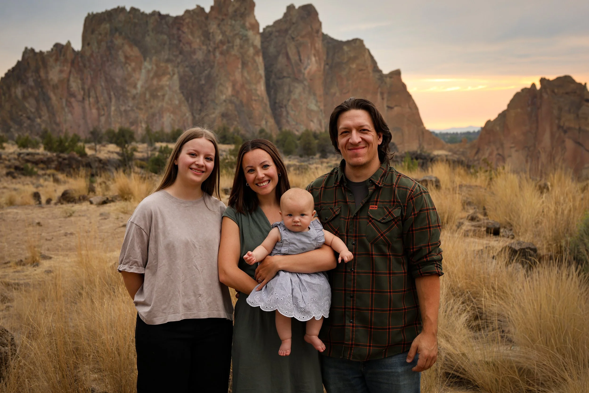A family of four standing outdoors at sunset in front of rocky cliffs and grassy terrain. The family includes a woman, a man, a young girl, and a baby. The woman is smiling, the man is smiling, the young girl has a neutral expression, and the baby is looking directly at the camera.