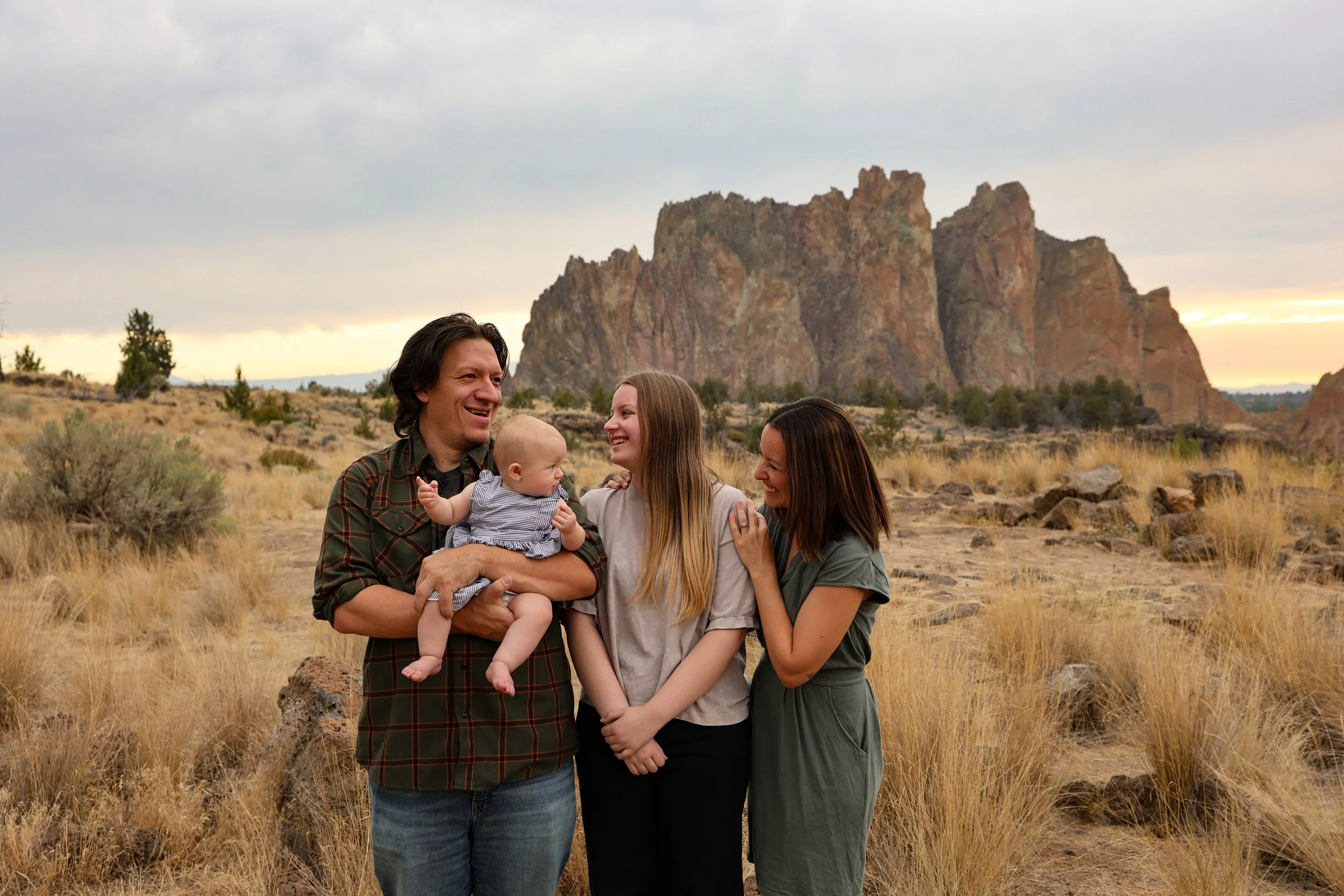 A family of four standing outdoors in a desert landscape with large rock formations in the background, smiling and interacting with each other.