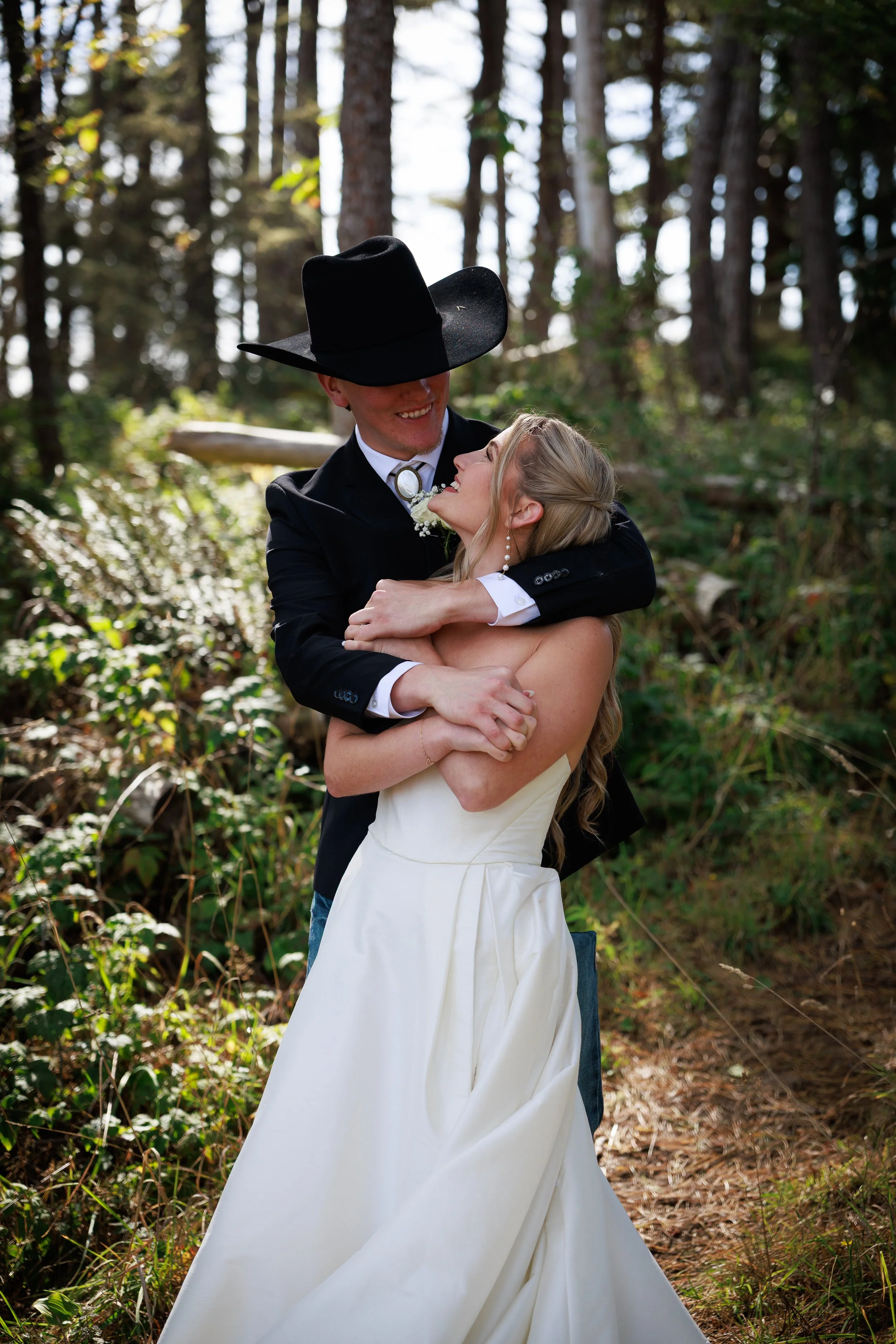 A couple dressed in wedding attire, embracing in a forest with trees and greenery in the background.