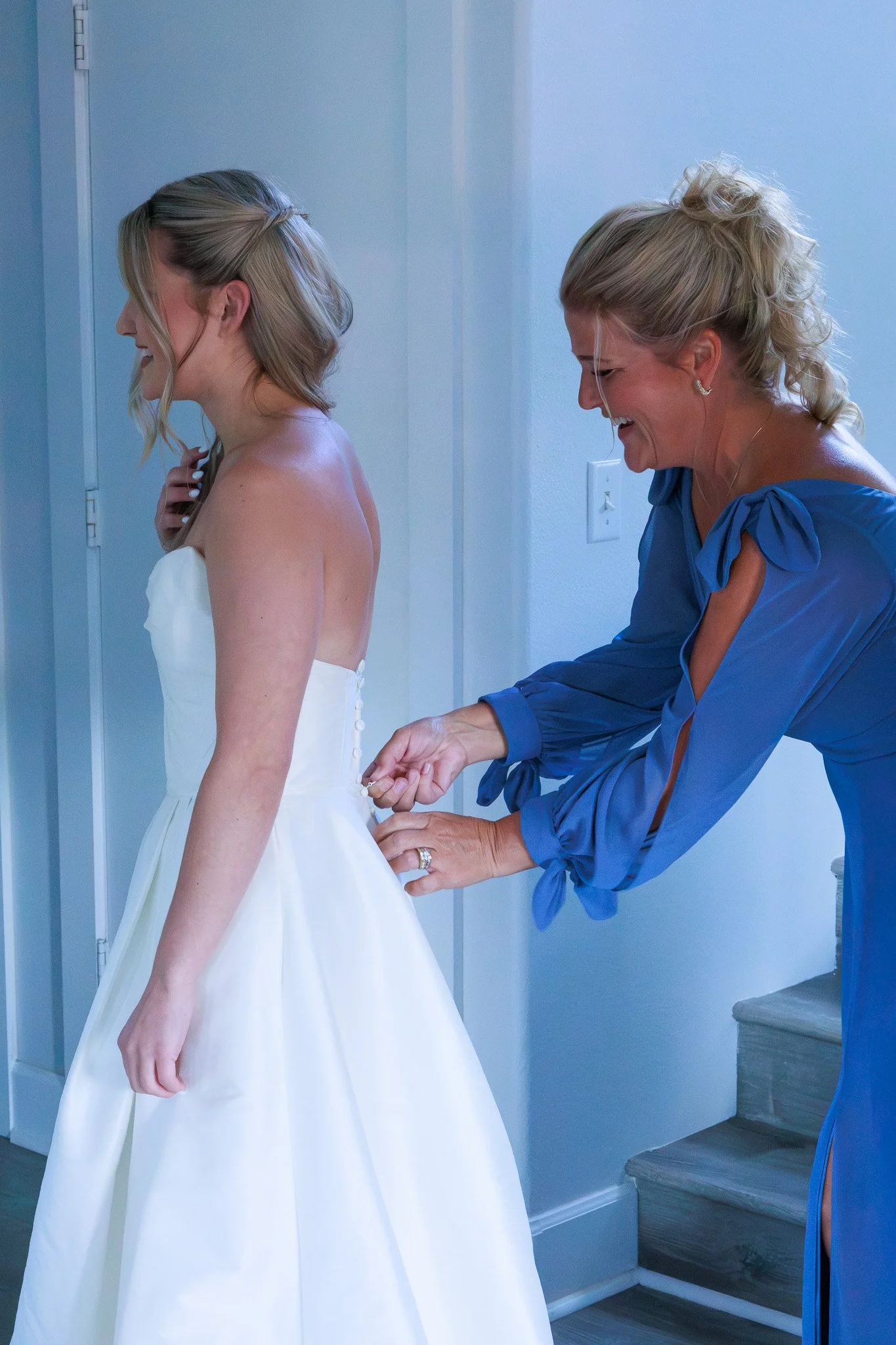 A woman in a blue dress is helping a bride put on buttons on the back of her white wedding dress.