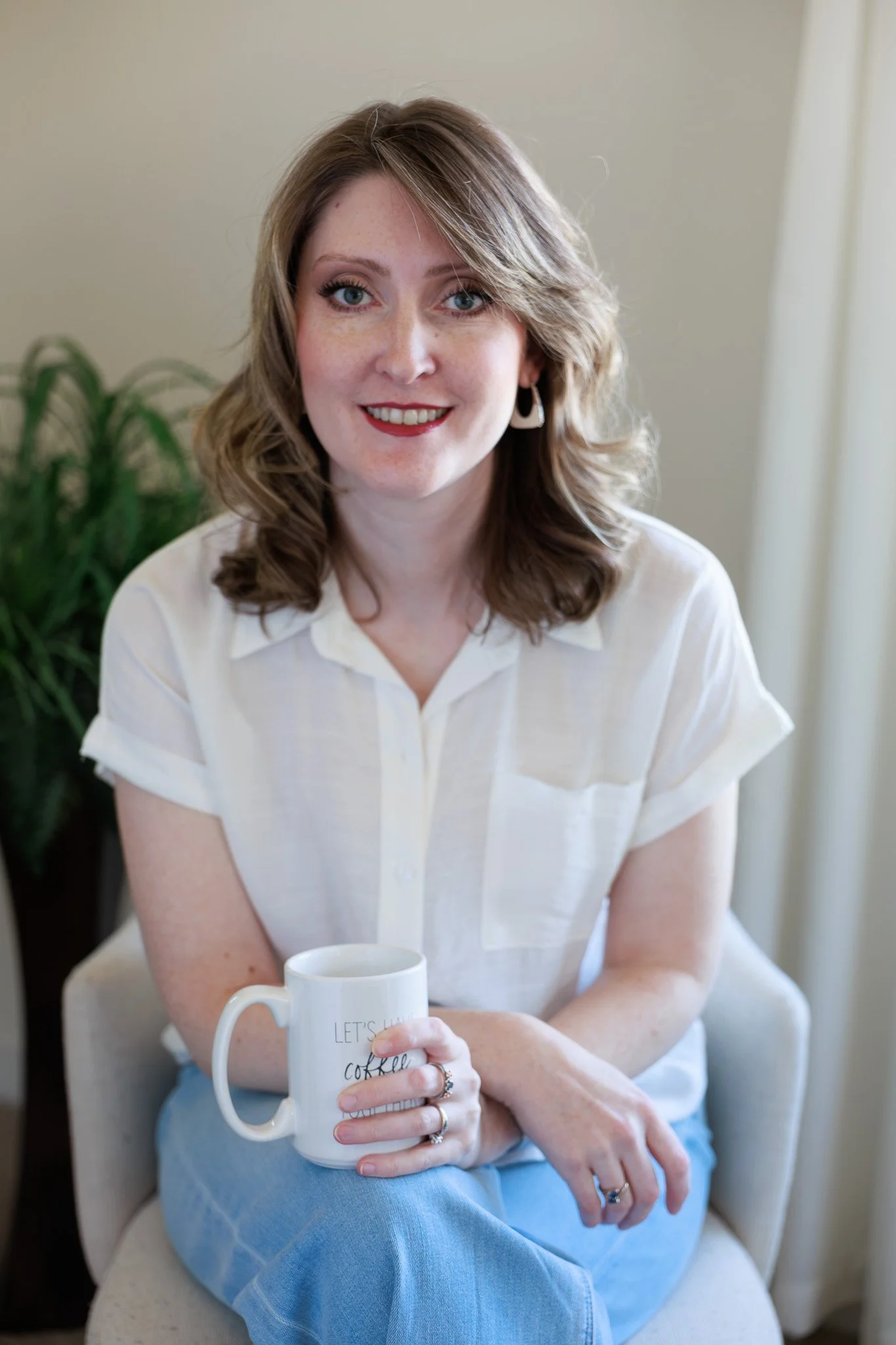 A woman with wavy brown hair and blue eyes sitting on a cream-colored armchair, holding a white mug with the words "Let's Have Coffee" written on it. She is wearing a white short-sleeved blouse and earrings, with a slight smile, in a room with a green plant in the background and light-colored curtains.