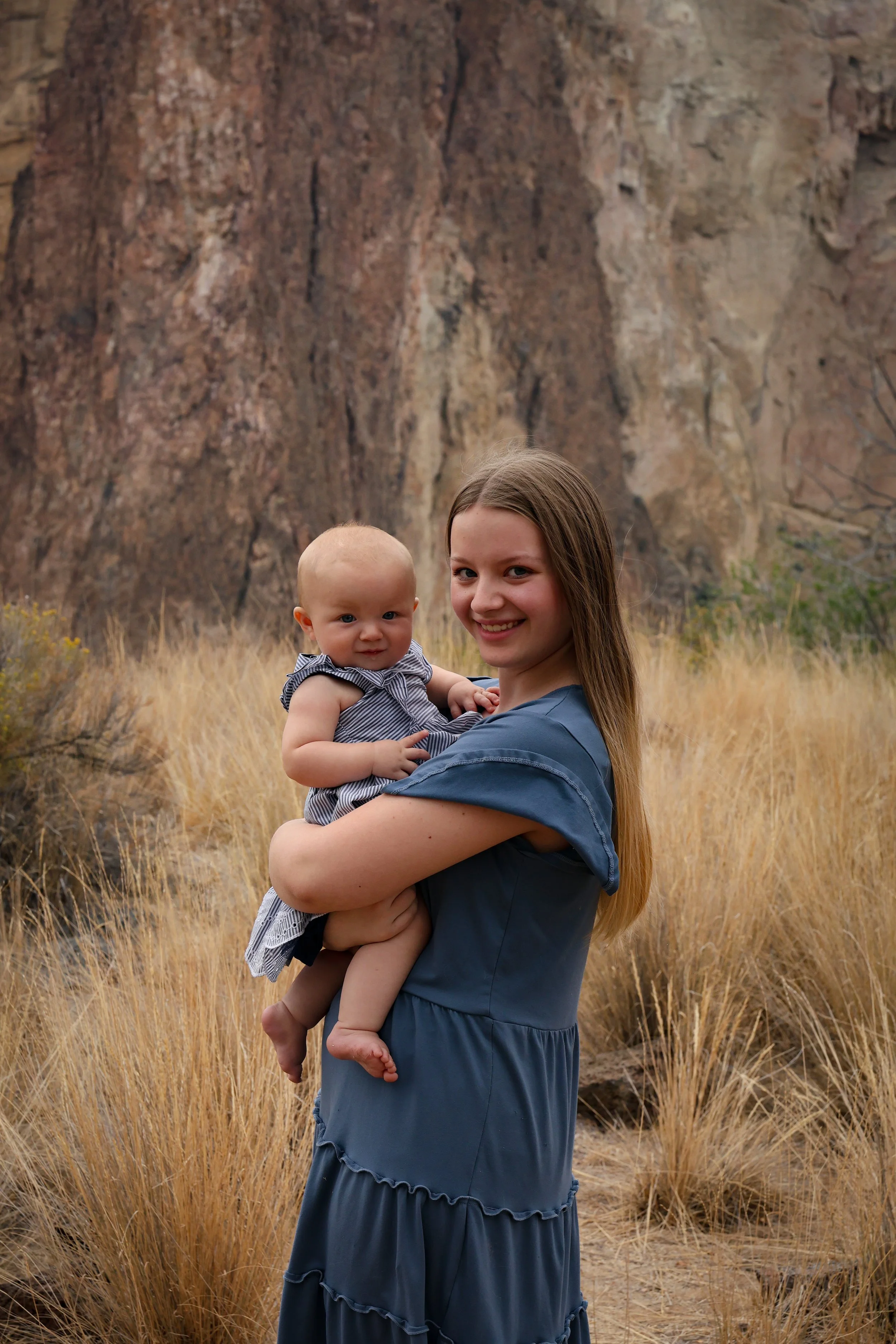 A woman holding a baby in a field with tall yellow grass and a large rocky cliff in the background.