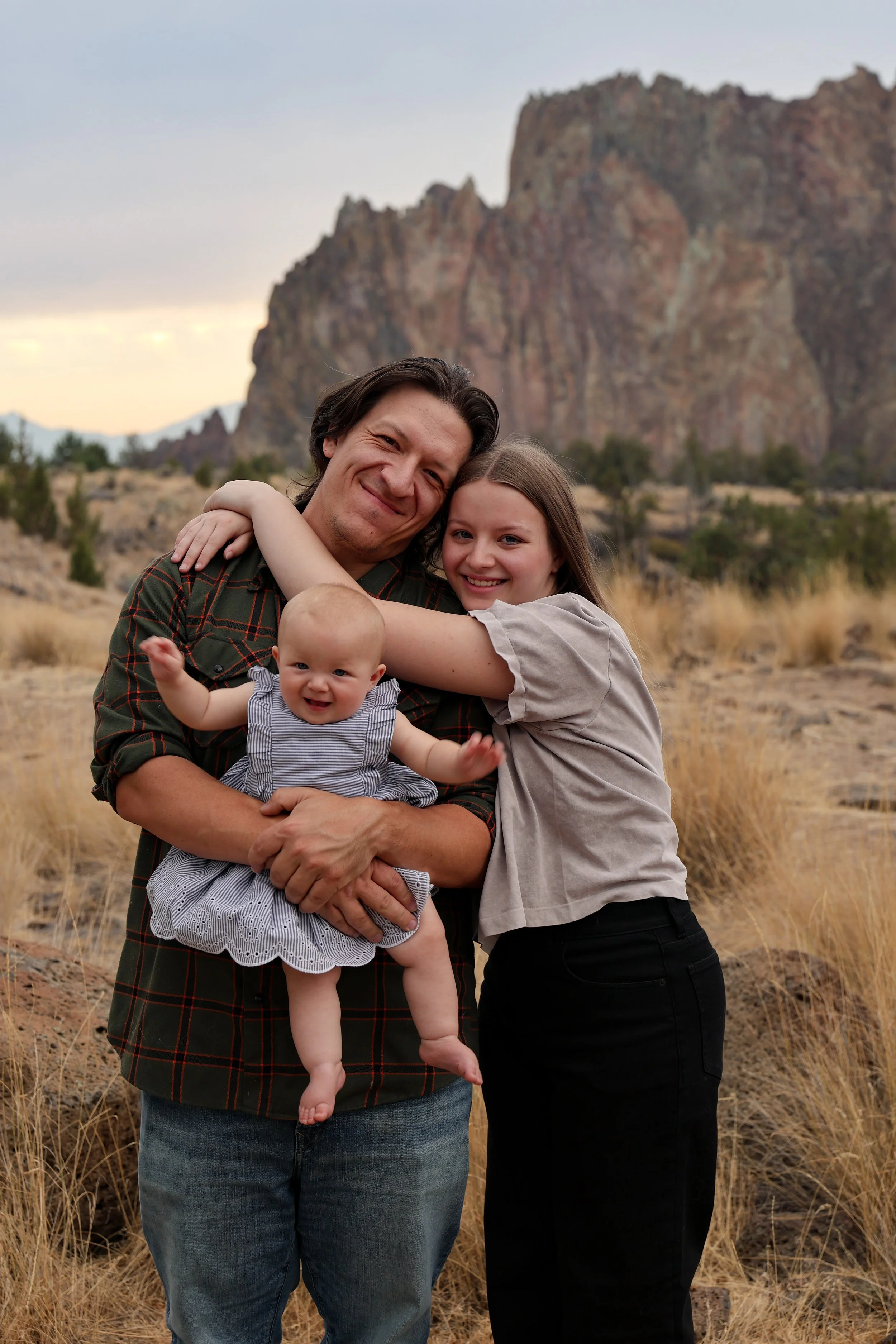 A family of four stands outdoors in front of a large rocky mountain. The father, mother, and two children are happily smiling, with the father holding the younger child. The older girl hugs the father, and the younger child is in the father's arms.