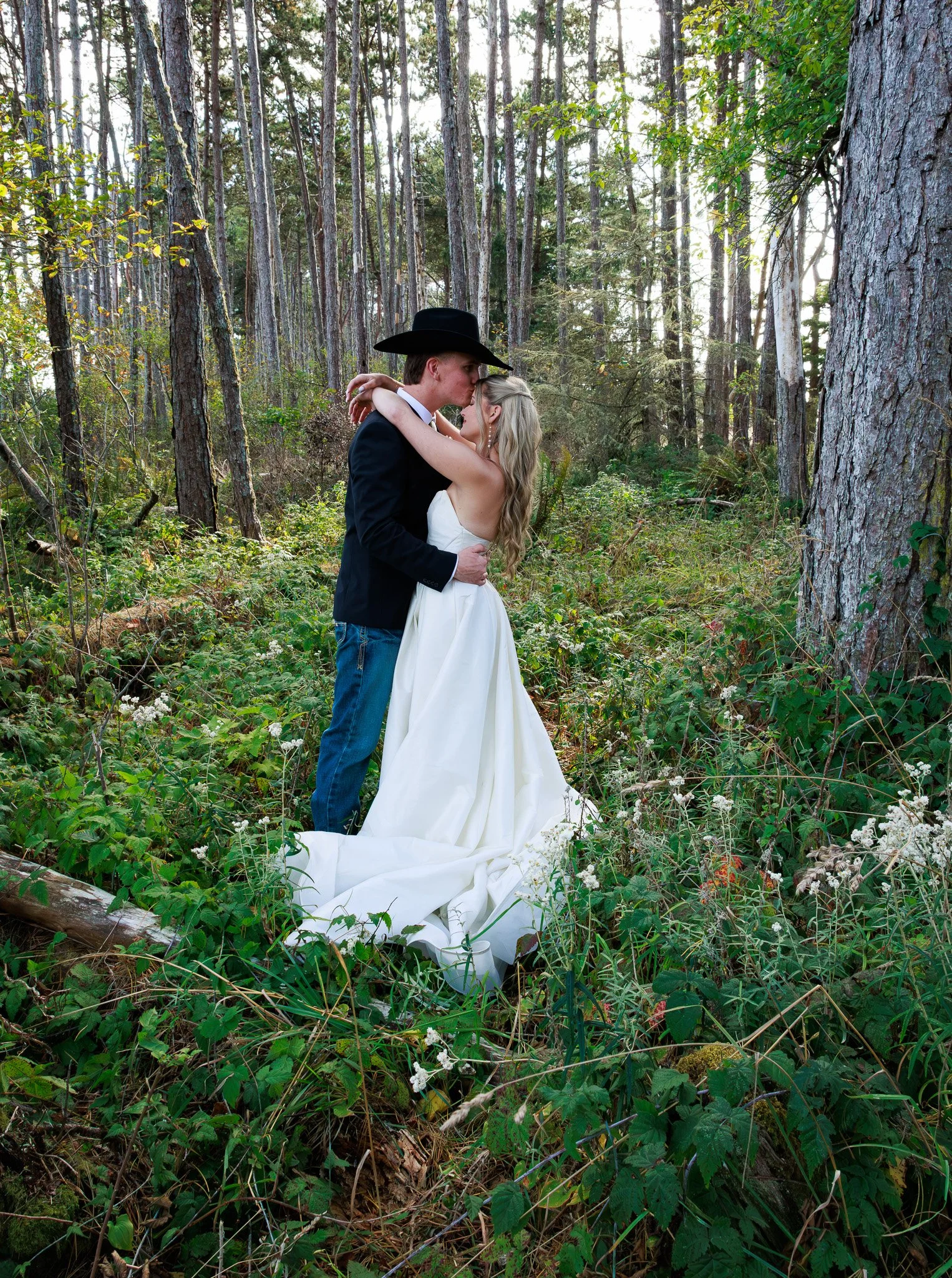 A couple dressed in wedding attire, with the groom wearing a black cowboy hat and the bride in a white gown, sharing an intimate moment in a dense forest.