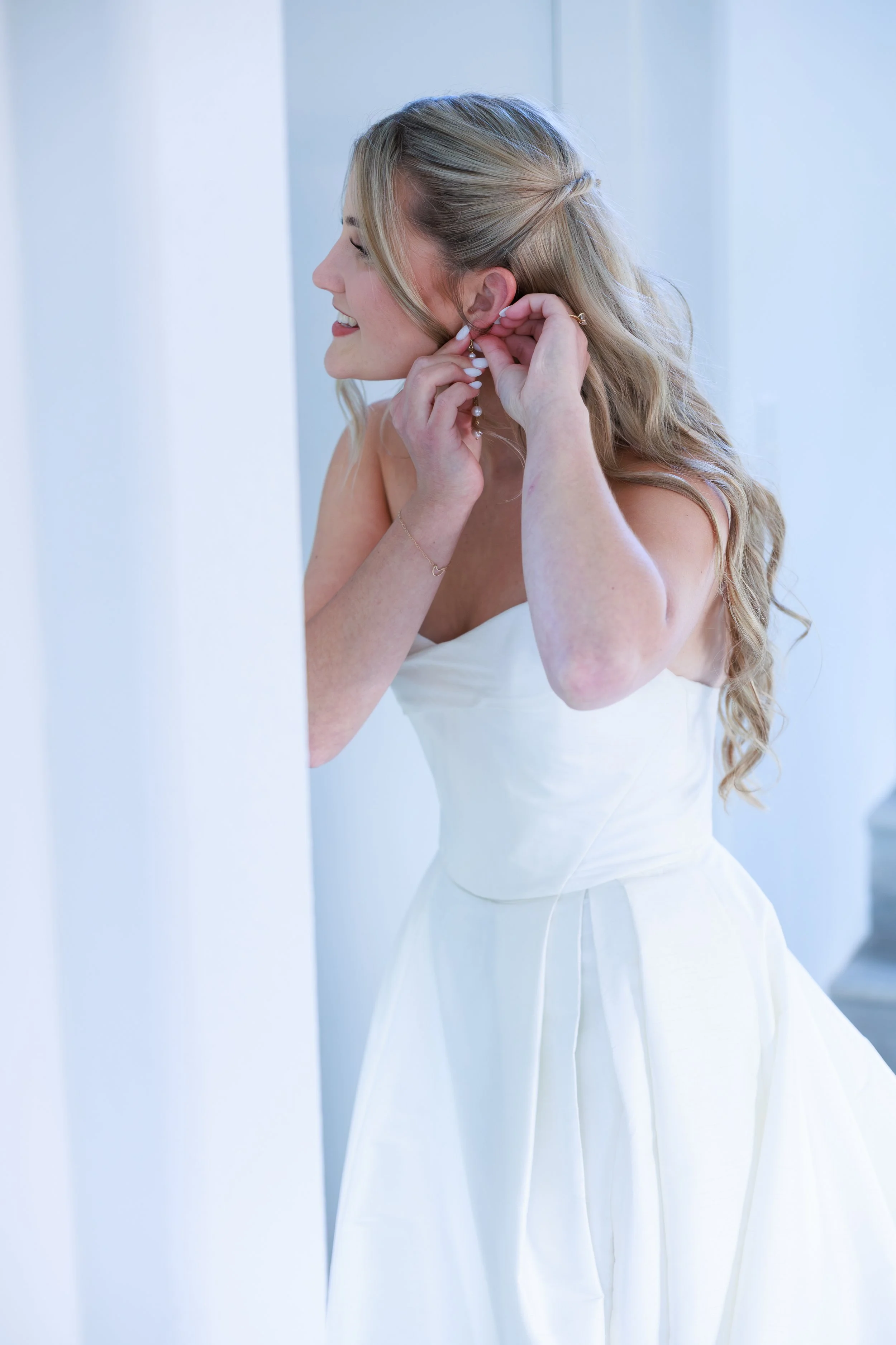 A woman with long, blonde hair styled in loose curls, wearing a strapless white dress, is putting on an earring in a bright, white room.