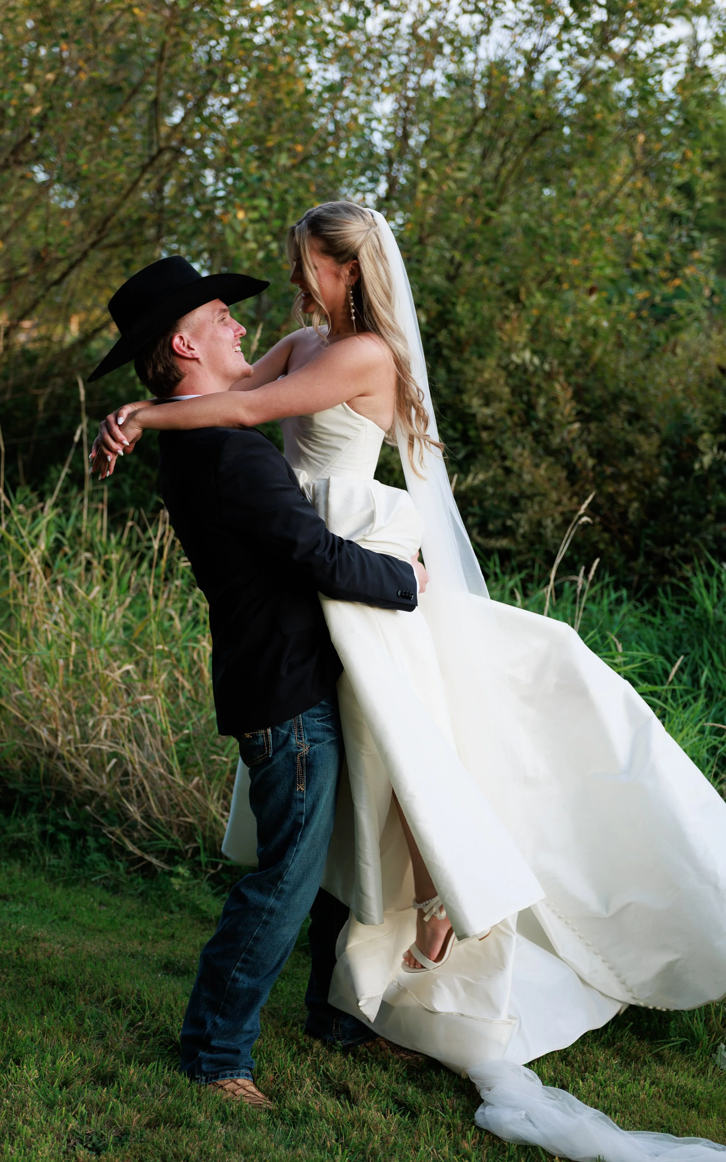 A groom lifting a bride outdoors with green trees and grass, both smiling and looking at each other.