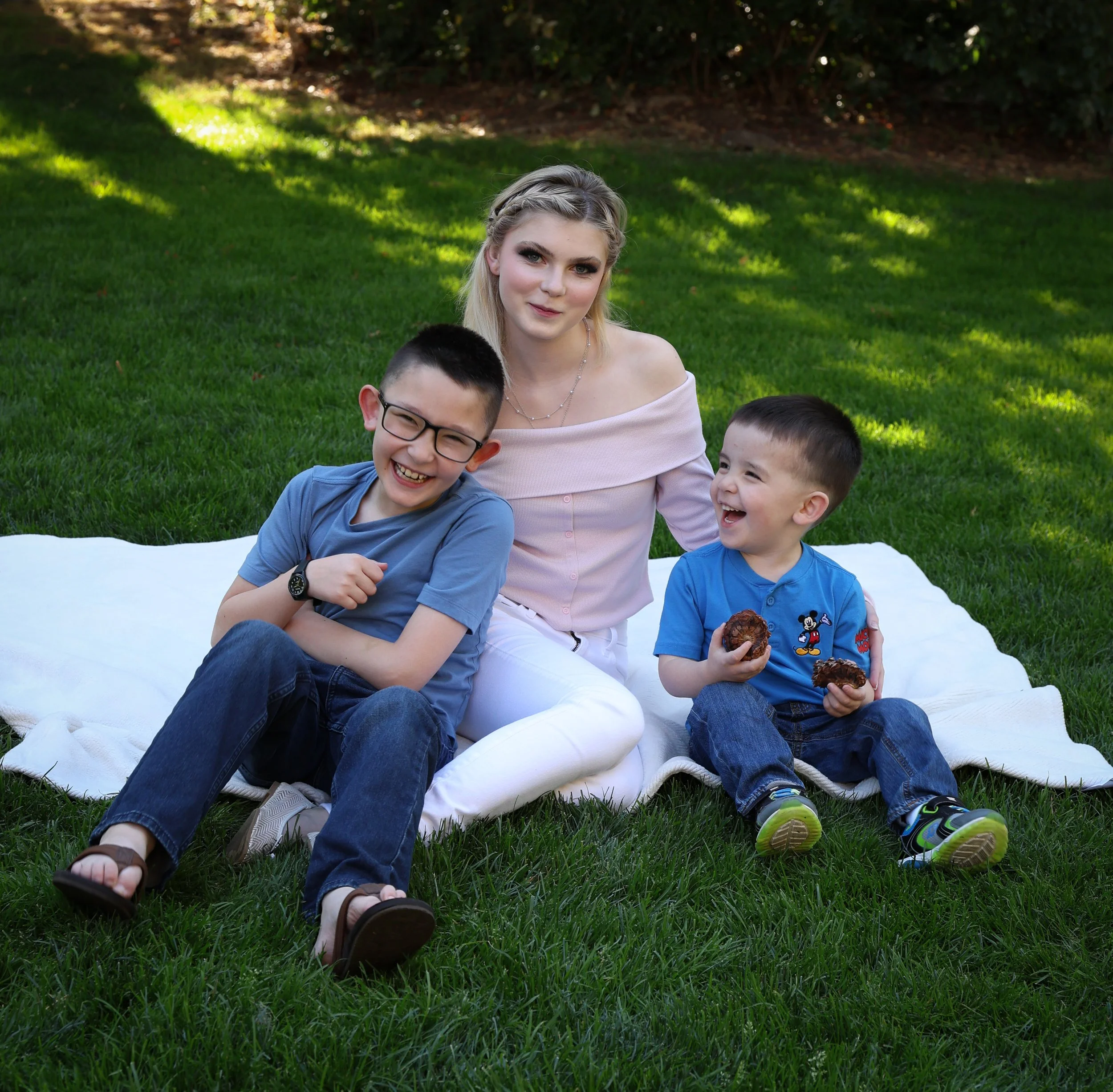 A woman with blonde hair and three children sitting on a white blanket on a grassy lawn, smiling, with trees and sunlight in the background.