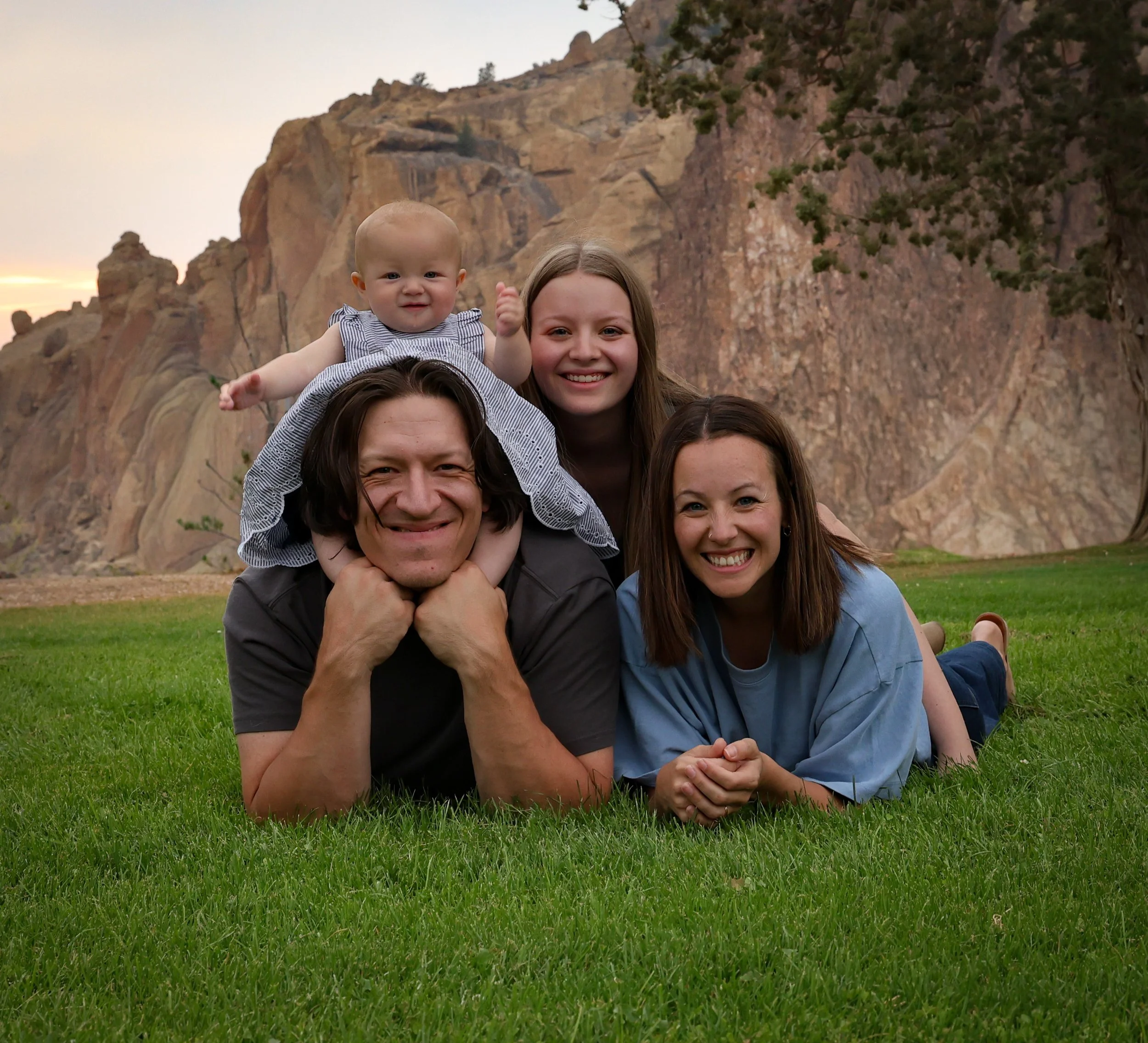 A smiling man and woman lie on green grass with a young girl and a baby girl on top of the man's back, set against a backdrop of rocky cliffs and a tree at dusk.