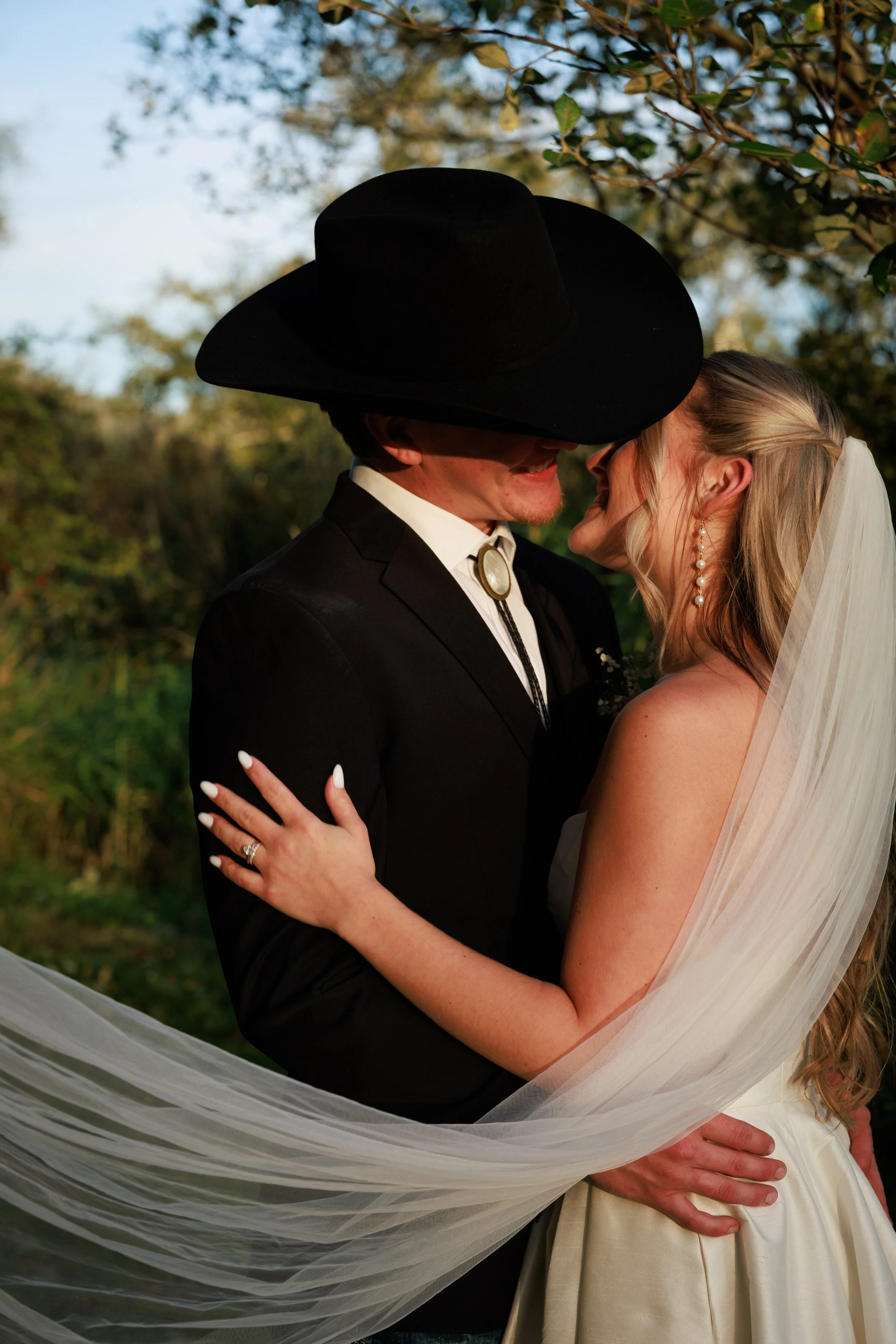 A bride and groom embrace outdoors, with the groom wearing a black hat and suit, and the bride in a wedding dress with pearl earrings, standing in front of trees during sunset.