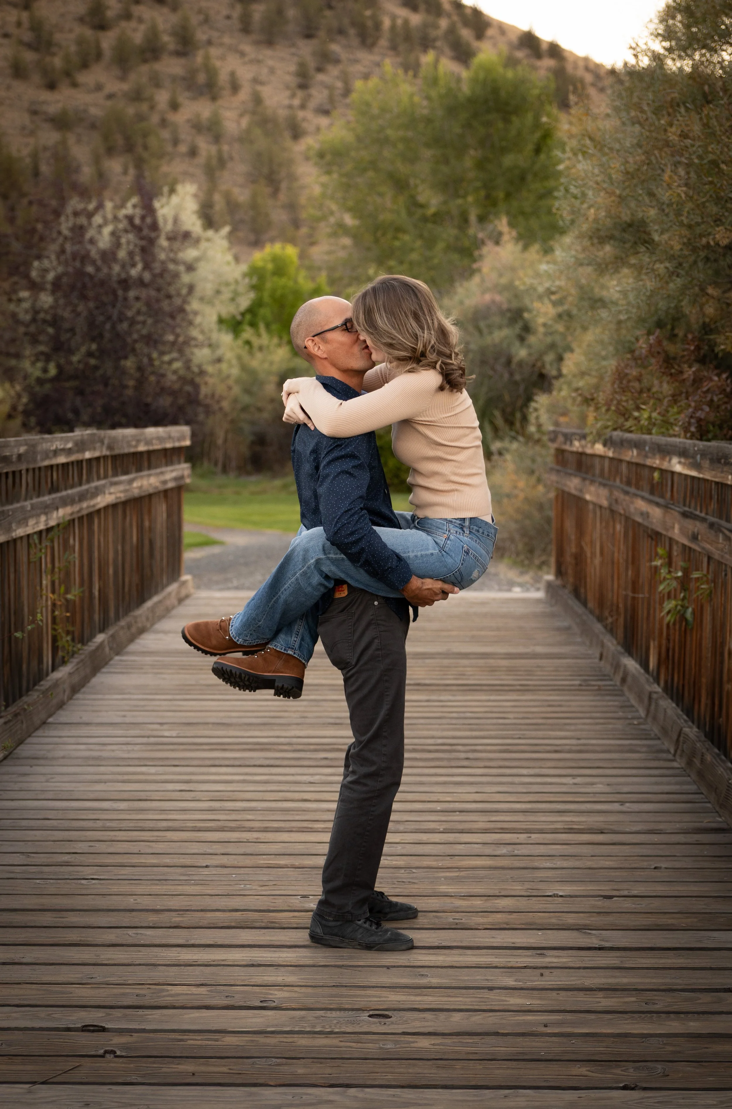 A man holding a woman in his arms and kissing her on the lips on a wooden bridge outdoors surrounded by trees and mountains.