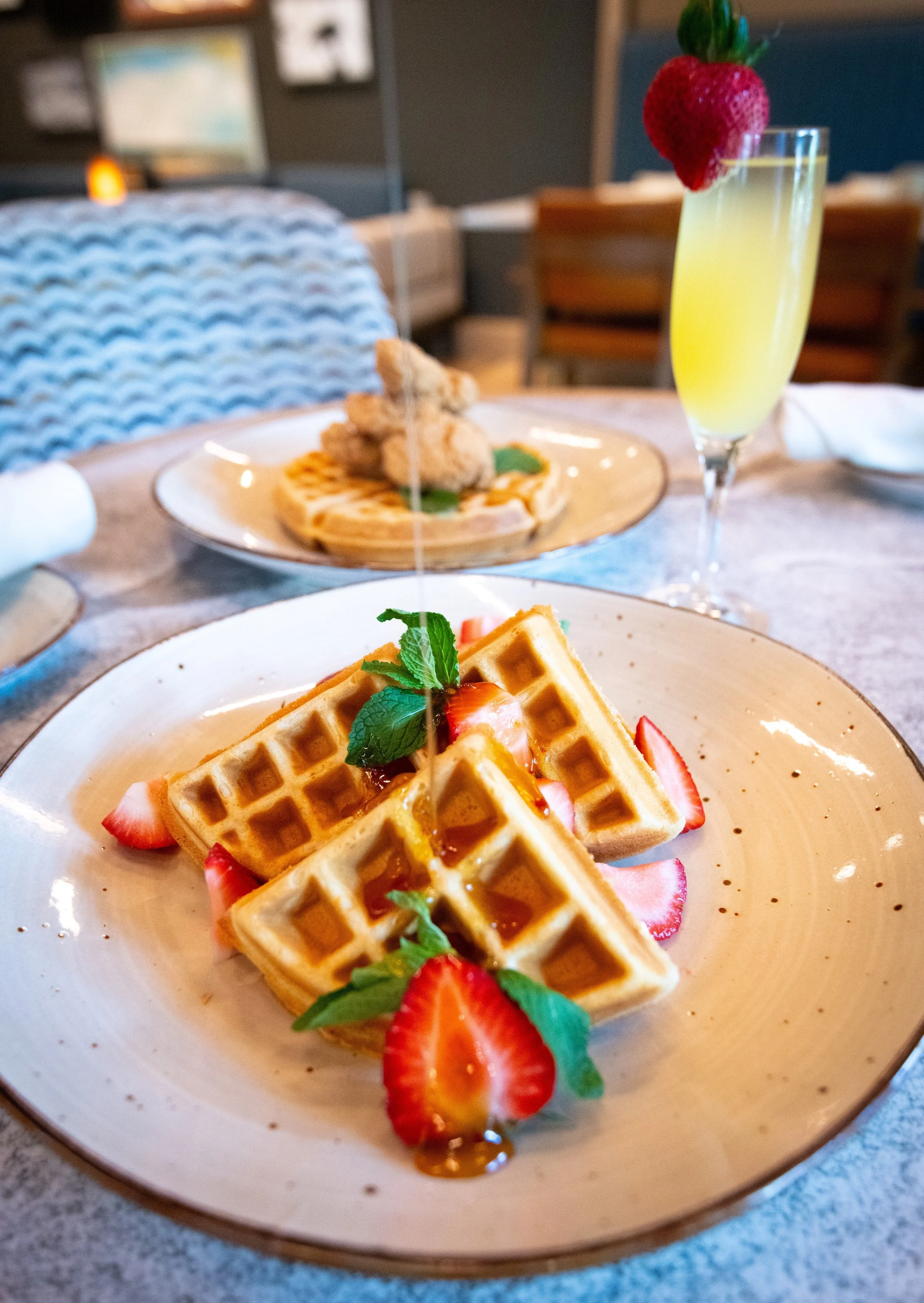A plate of waffles topped with strawberries and mint leaves in the foreground, a plate with waffles and a fried chicken slice in the background, and a glass of yellow-colored drink with a strawberry garnish in a restaurant setting.