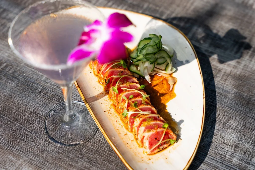 A plate with sliced tuna, garnished with microgreens, accompanied by cucumber salad, and a purple orchid flower, with a glass of cocktail garnished with a purple orchid on a wooden surface.