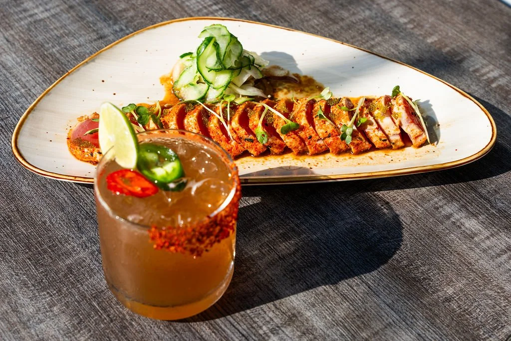 Sliced spicy tuna sushi roll garnished with microgreens, served on a long oval white plate with sliced cucumber and ginger, accompanied by a cocktail with lime, chili pepper, and salted rim on a dark wood table.