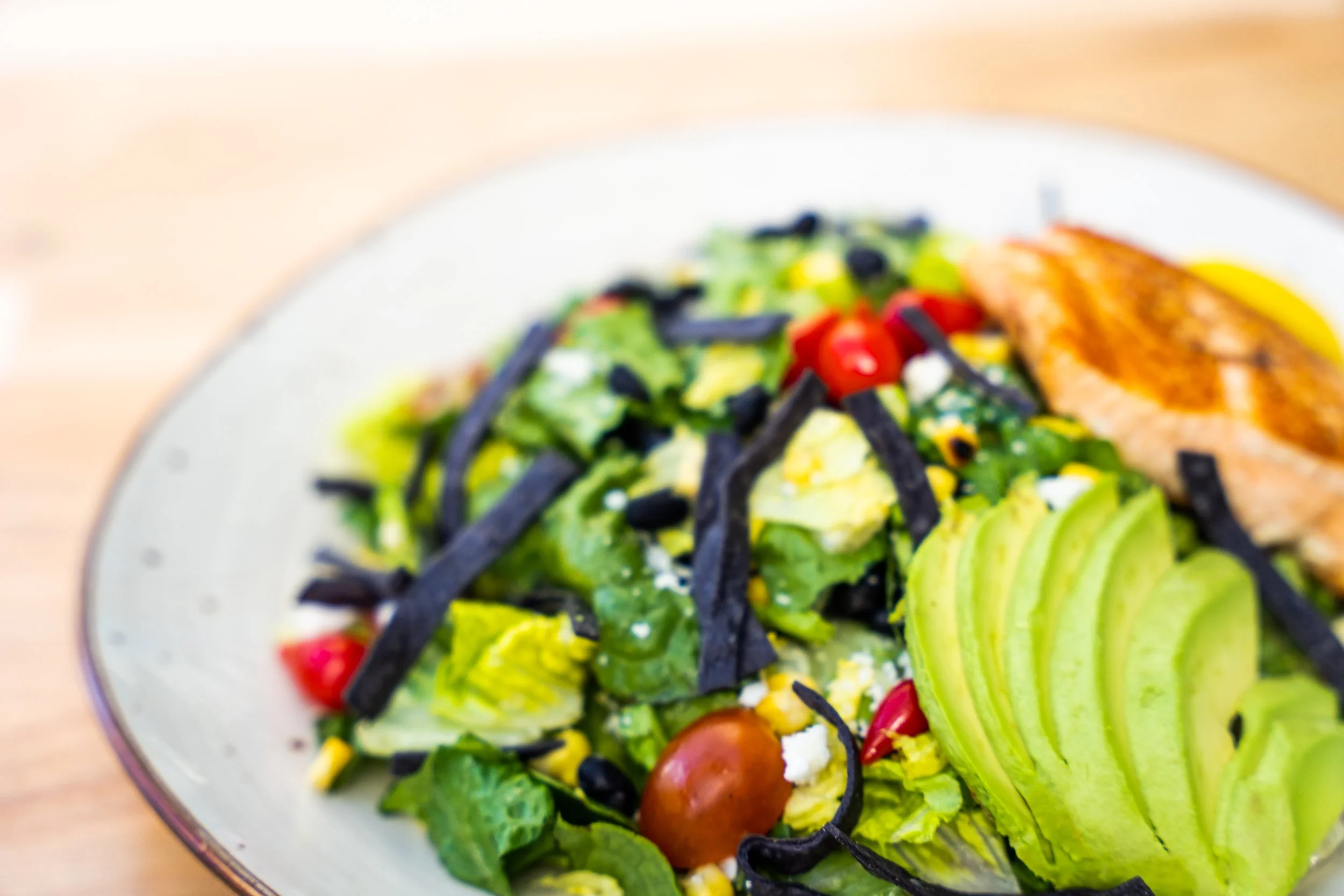A colorful salad on a white plate, featuring sliced avocado, cherry tomatoes, black beans, corn, and shredded cheese, with grilled chicken on the side.