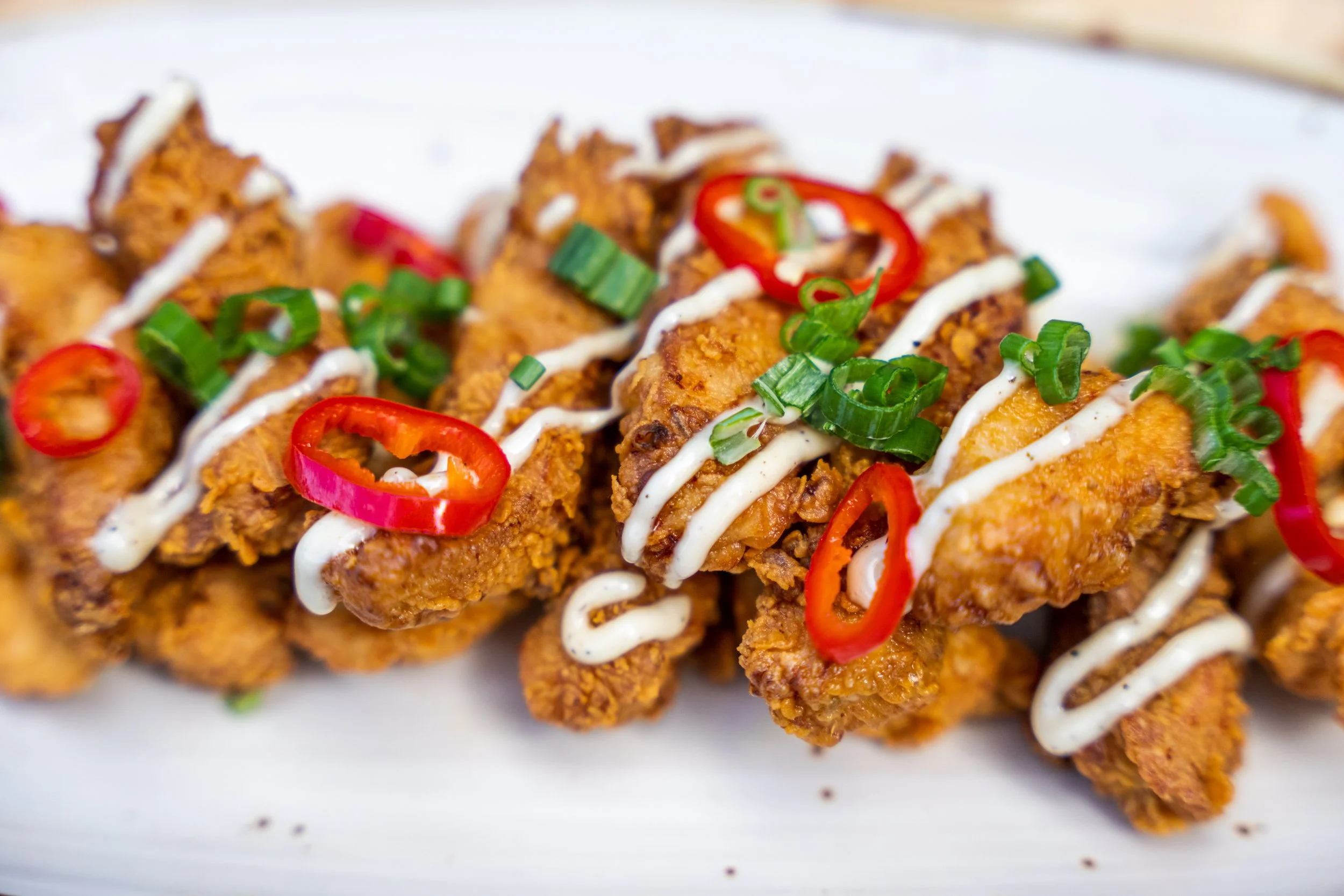 Close-up of crispy fried chicken pieces garnished with chopped green onions and sliced red chili peppers, drizzled with white and brown sauces.