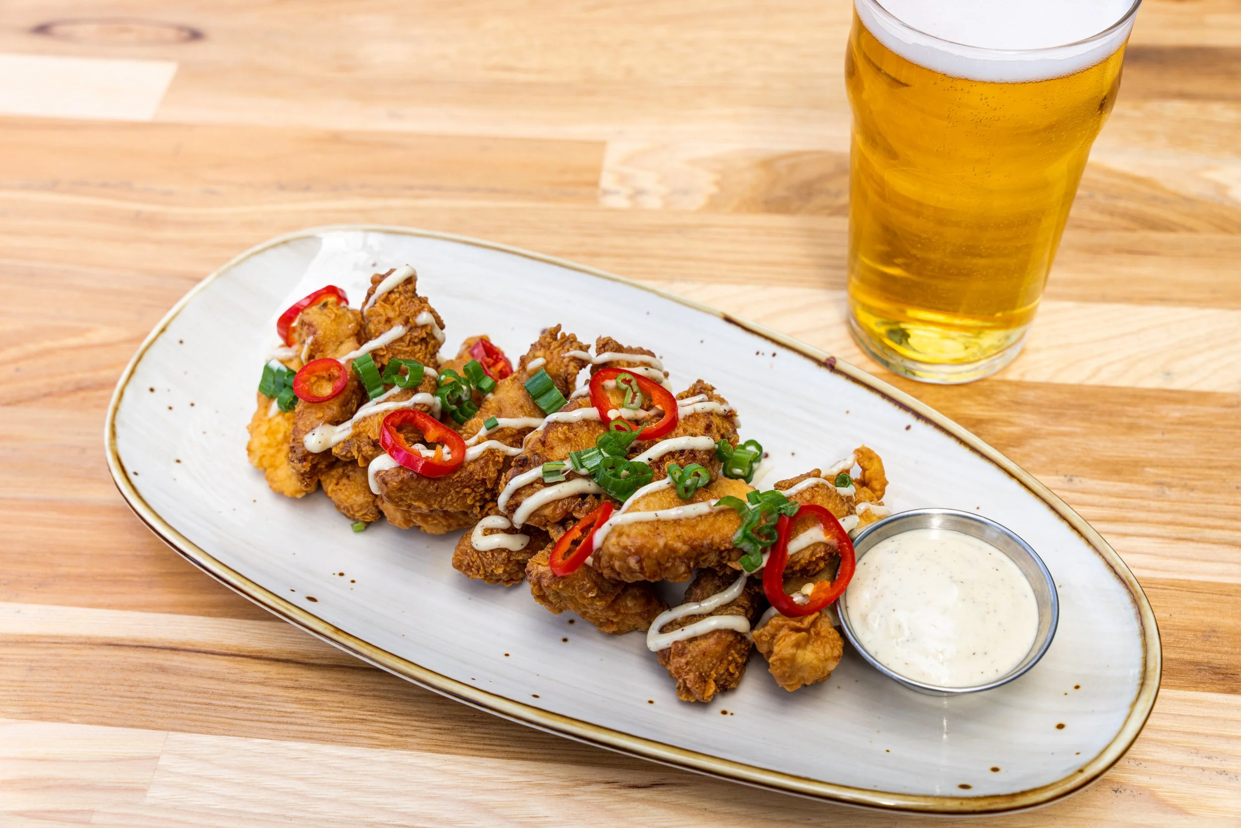 Fried chicken wings topped with sliced red chili peppers and chopped green onions, drizzled with white sauce, served on a white oval plate with a side of dipping sauce, with a glass of beer in the background on a wooden table.