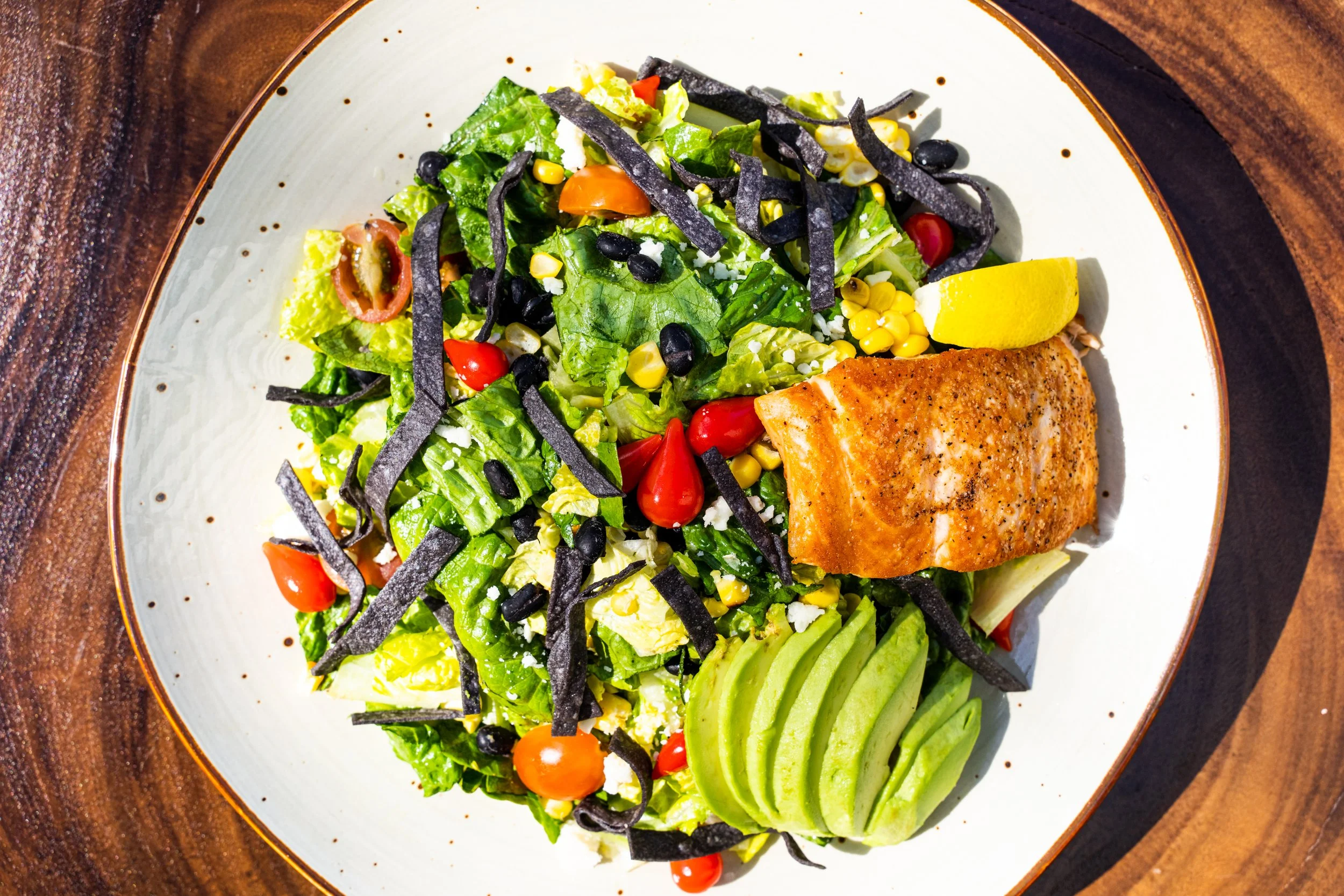 A plate of salad with lettuce, cherry tomatoes, corn, black beans, avocado slices, lemon wedge, and pieces of grilled salmon.