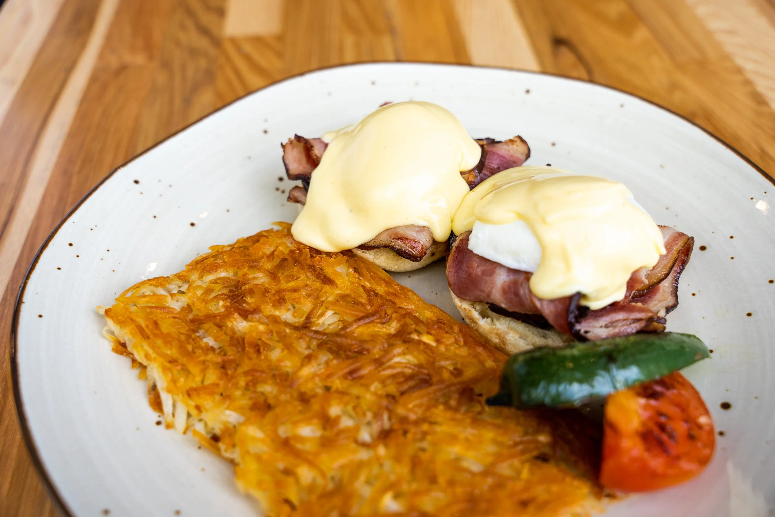 Dish with eggs Benedict topped with hollandaise sauce, hash browns, grilled vegetables, and bacon on a white plate with black speckles.