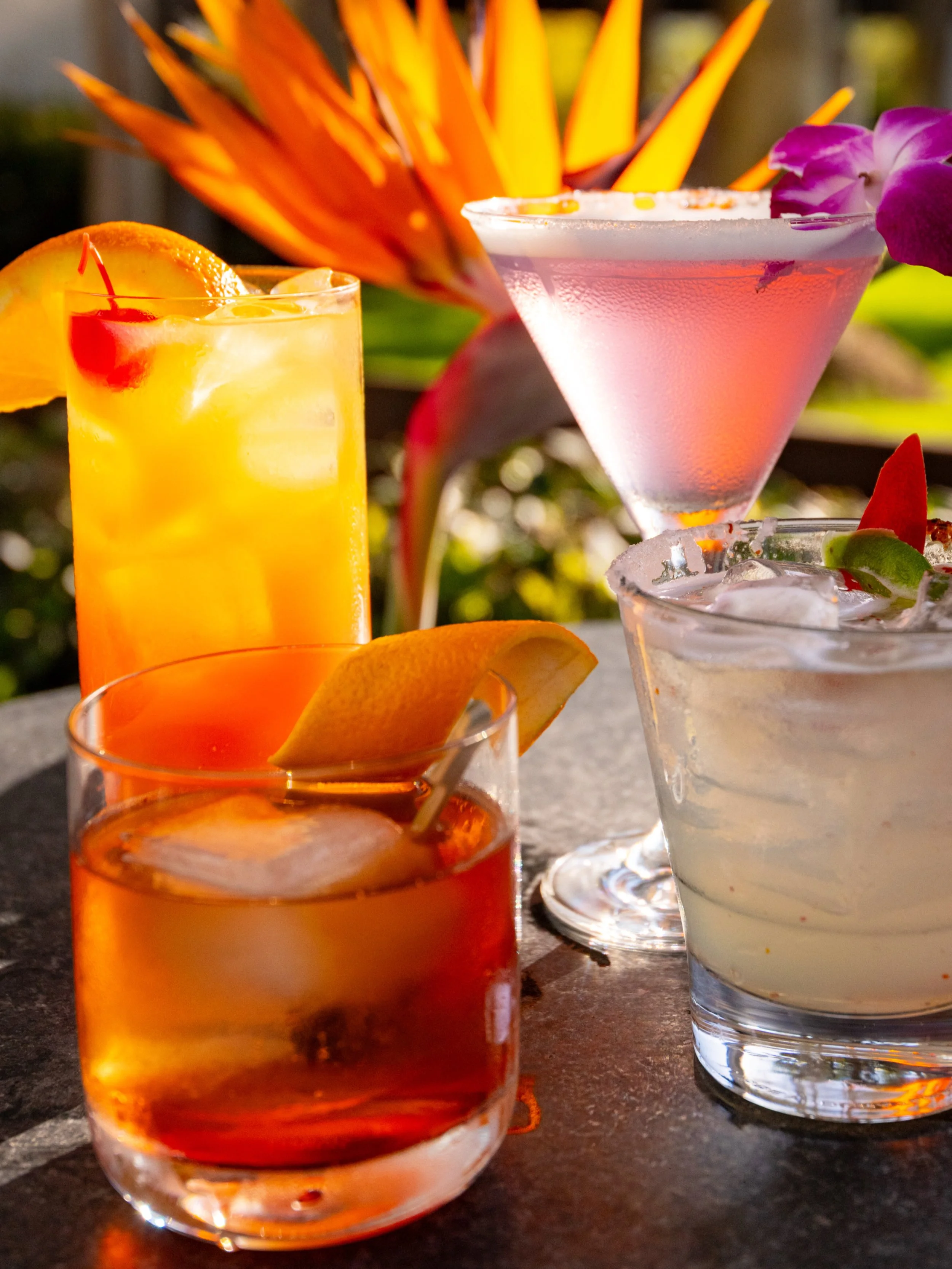 Various colorful cocktails with fruit garnishes on a dark table, including an orange-colored drink with an orange slice, a pink cocktail in a martini glass, and a clear drink with lime and cherry