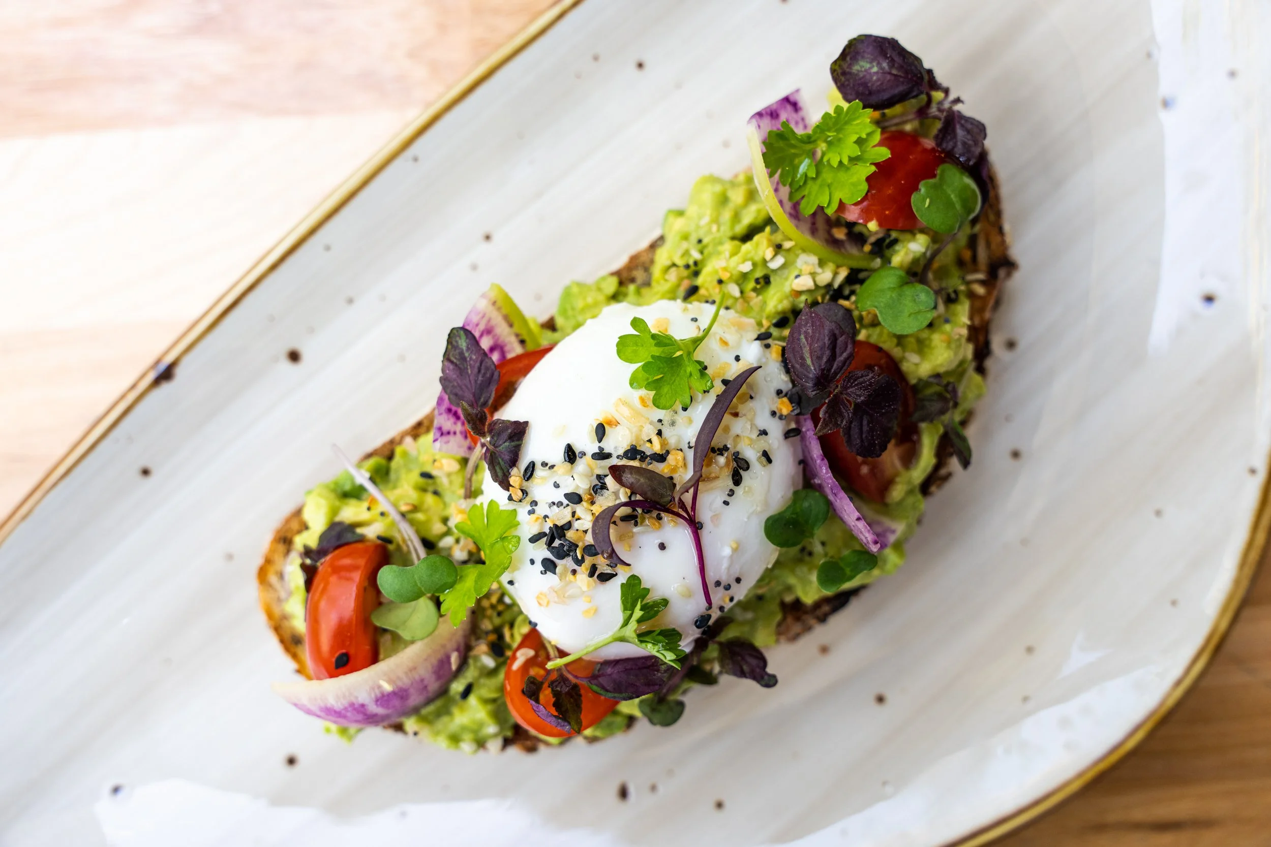 Close-up of avocado toast topped with cherry tomatoes, poached egg, microgreens, and sprinkled with sesame seeds and chopped nuts, served on a decorative plate.