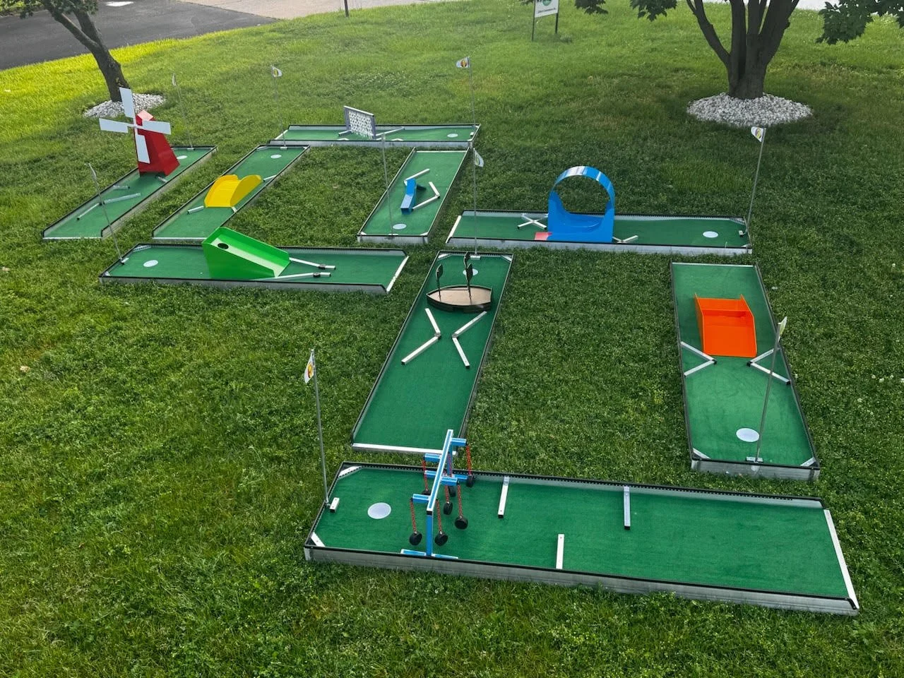 Miniature mini-golf course with various colorful obstacles and structures on a grassy area, including a windmill, a ramp, a curved tunnel, and small flags marking the holes, surrounded by trees and bushes.