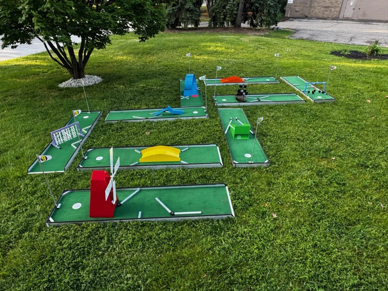 Miniature golf course set up on a grassy lawn with various obstacles, including a red obstacle with windmill blades, a yellow ramp, a green obstacle, and small flags on some holes.