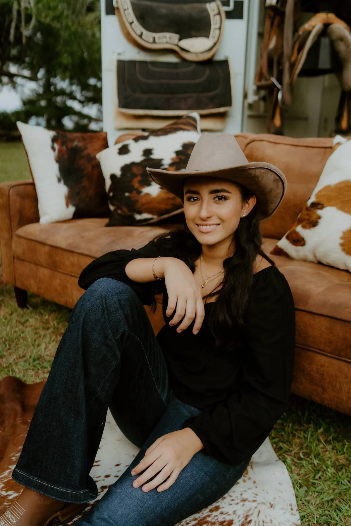 A young woman with dark hair, wearing a brown cowboy hat, black long-sleeve top, and blue jeans, sitting on a cowhide rug outside. Behind her is a leather sofa with cowhide pillows and a nature background with trees.