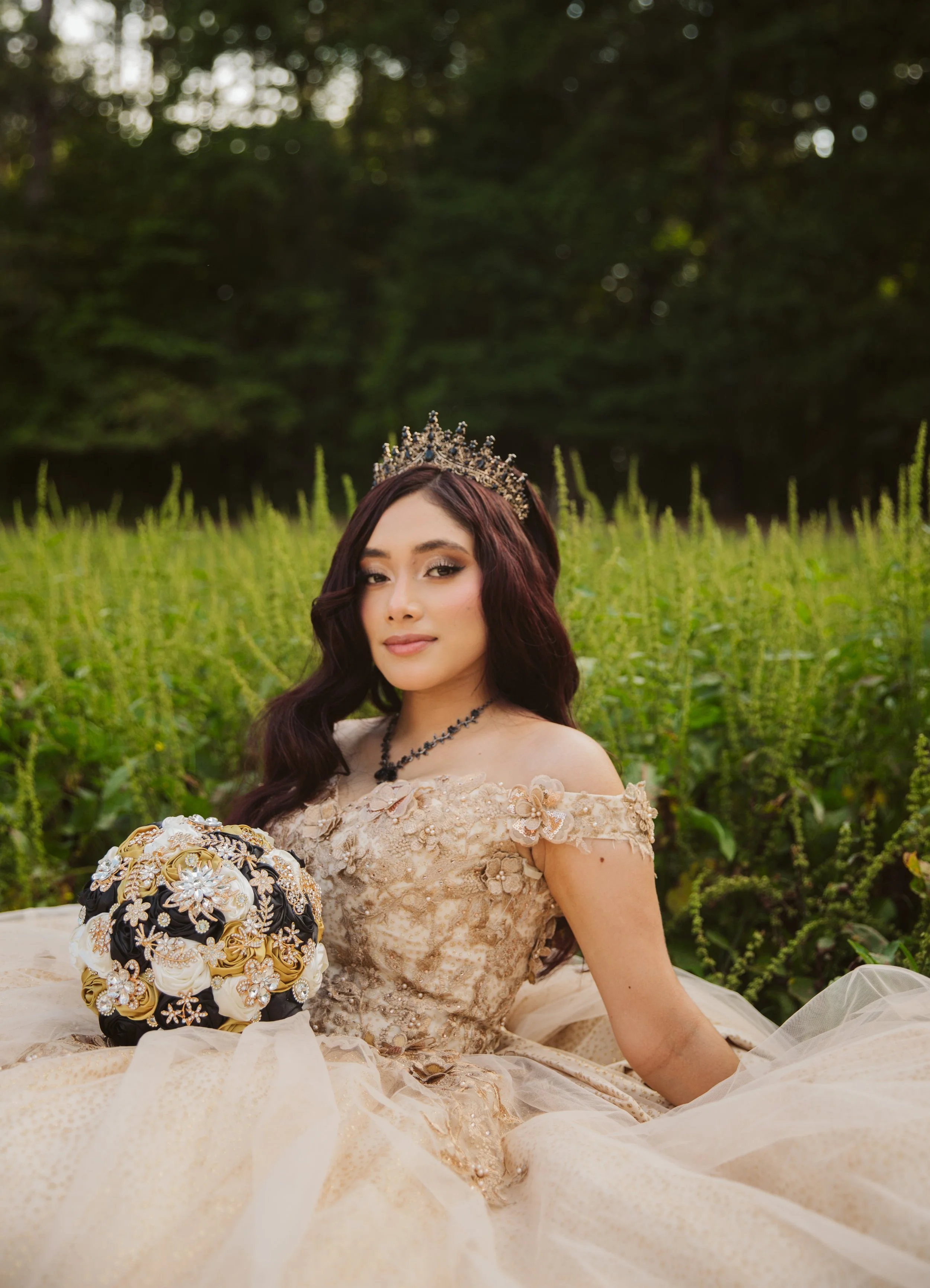 A woman in a beige, floral lace gown and a crown is sitting outdoors in a garden with greenery and tall plants, holding a bouquet of flowers.