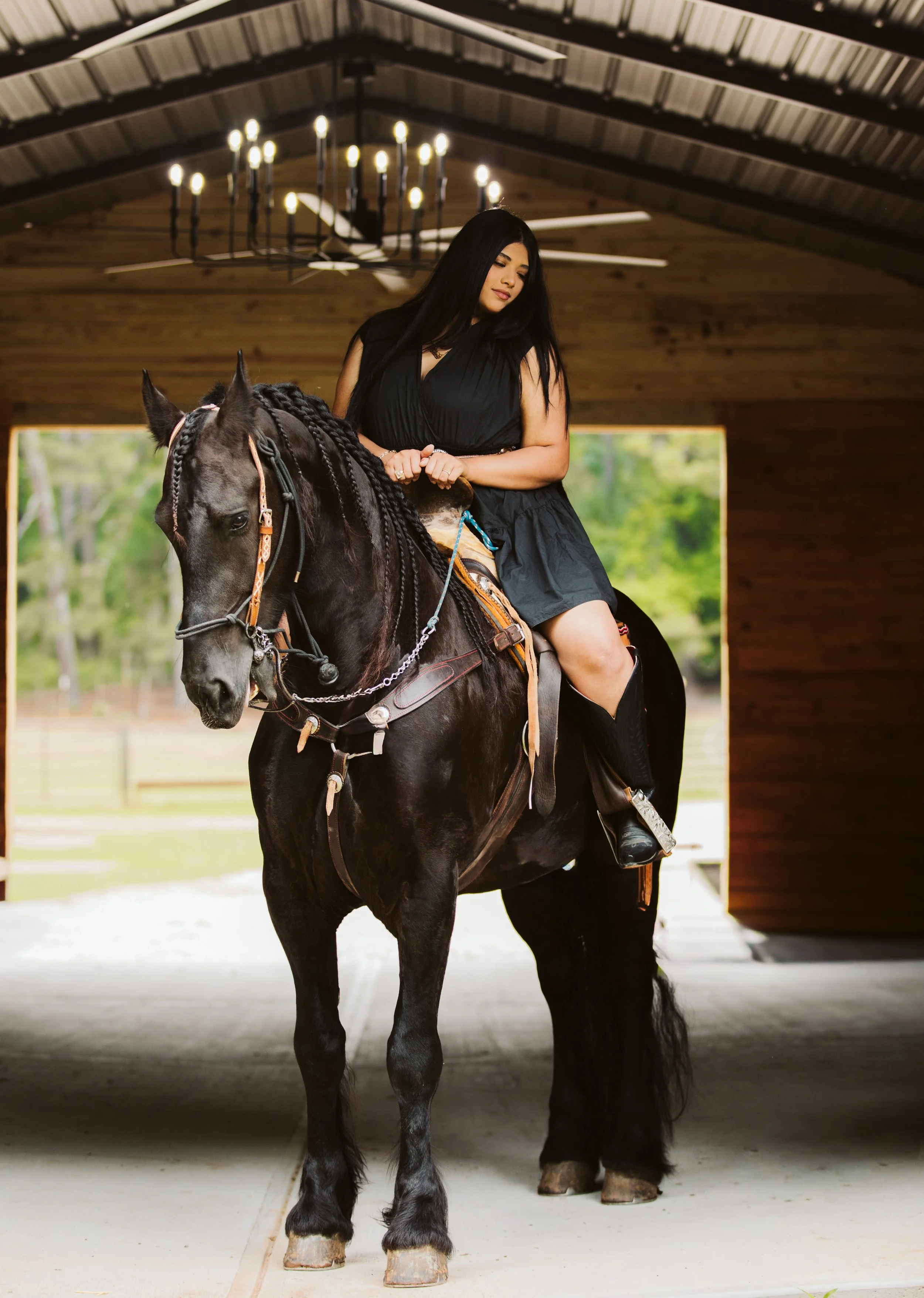 A woman riding a black horse inside a wooden structure with a chandelier overhead and large open windows showing green trees outside.