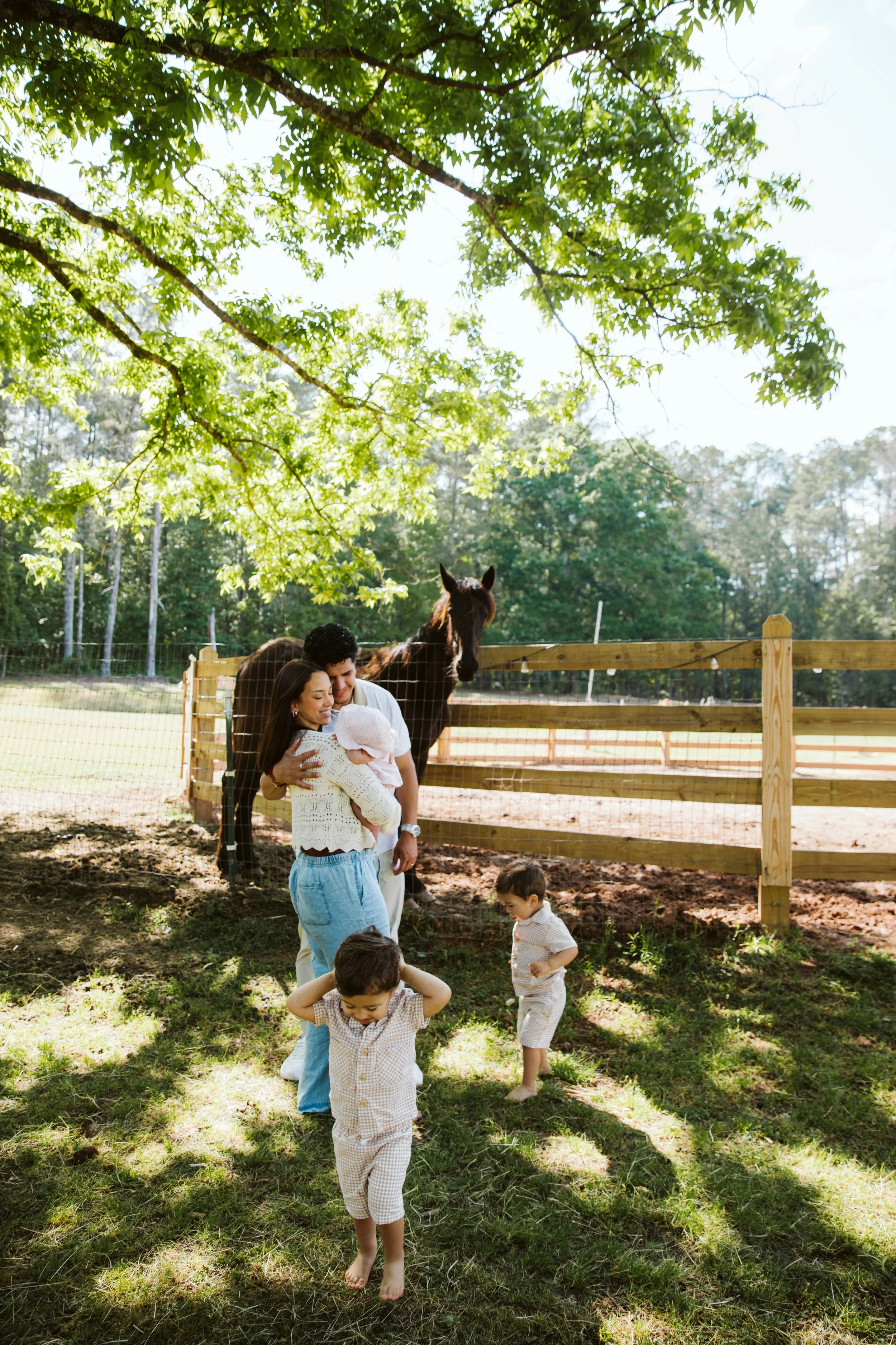 A family of five, including two young children, enjoying a sunny day outdoors near a wooden fence with a horse observing them behind the fence. The scene is set in a grassy area shaded by large leafy trees.