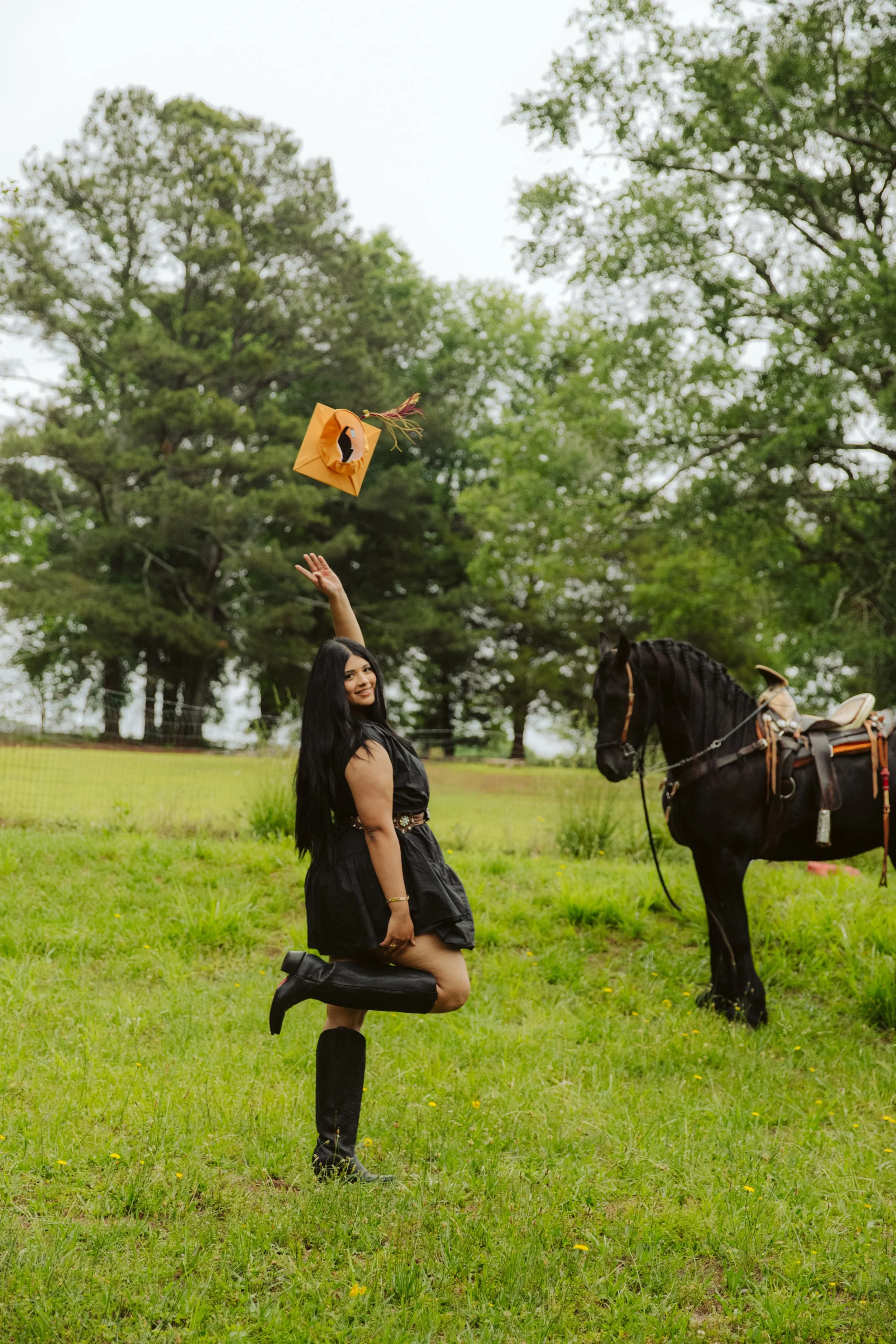 A woman in a black dress and boots kneeling on one knee in a grassy field, holding a hat in one hand, with a horse nearby and trees in the background.