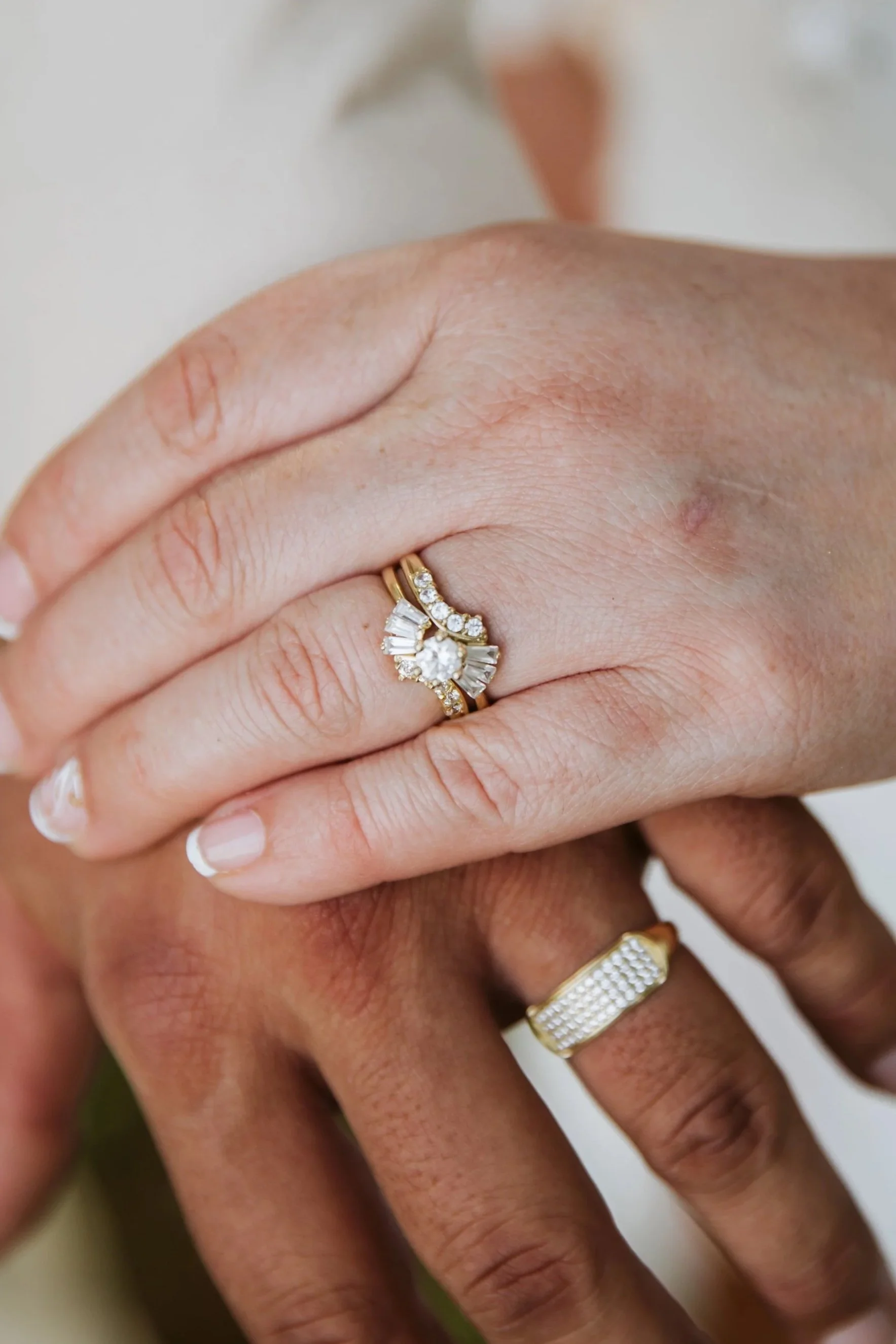 Close-up of hands showing multiple rings, including a prominent diamond ring on the ring finger with a gold band and a bundle of smaller diamonds, and a gold band with a textured pattern on the middle finger.