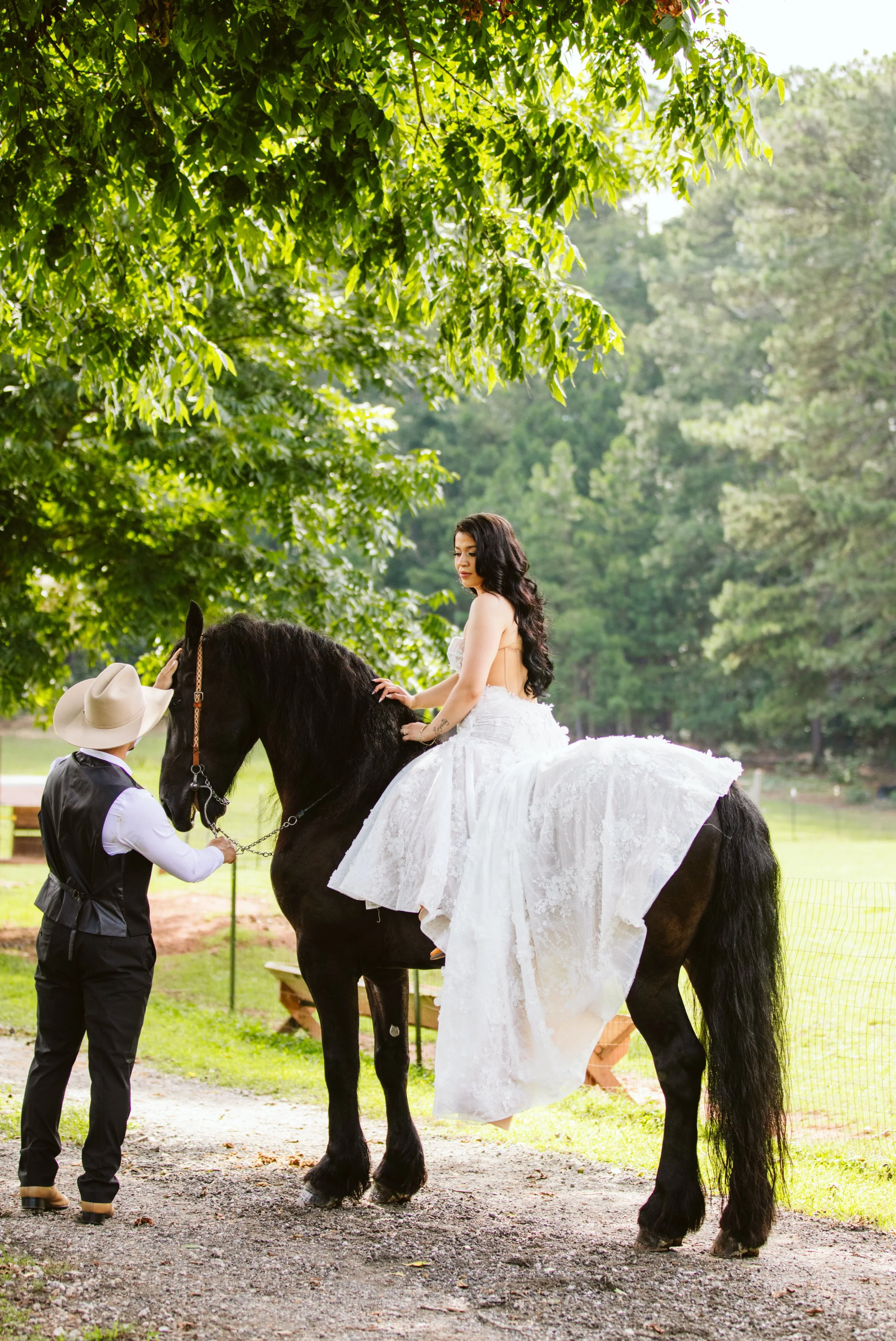 A woman in a wedding dress riding a black horse outdoors on a sunny day, with two men standing nearby, one holding the horse's reins, under green trees.