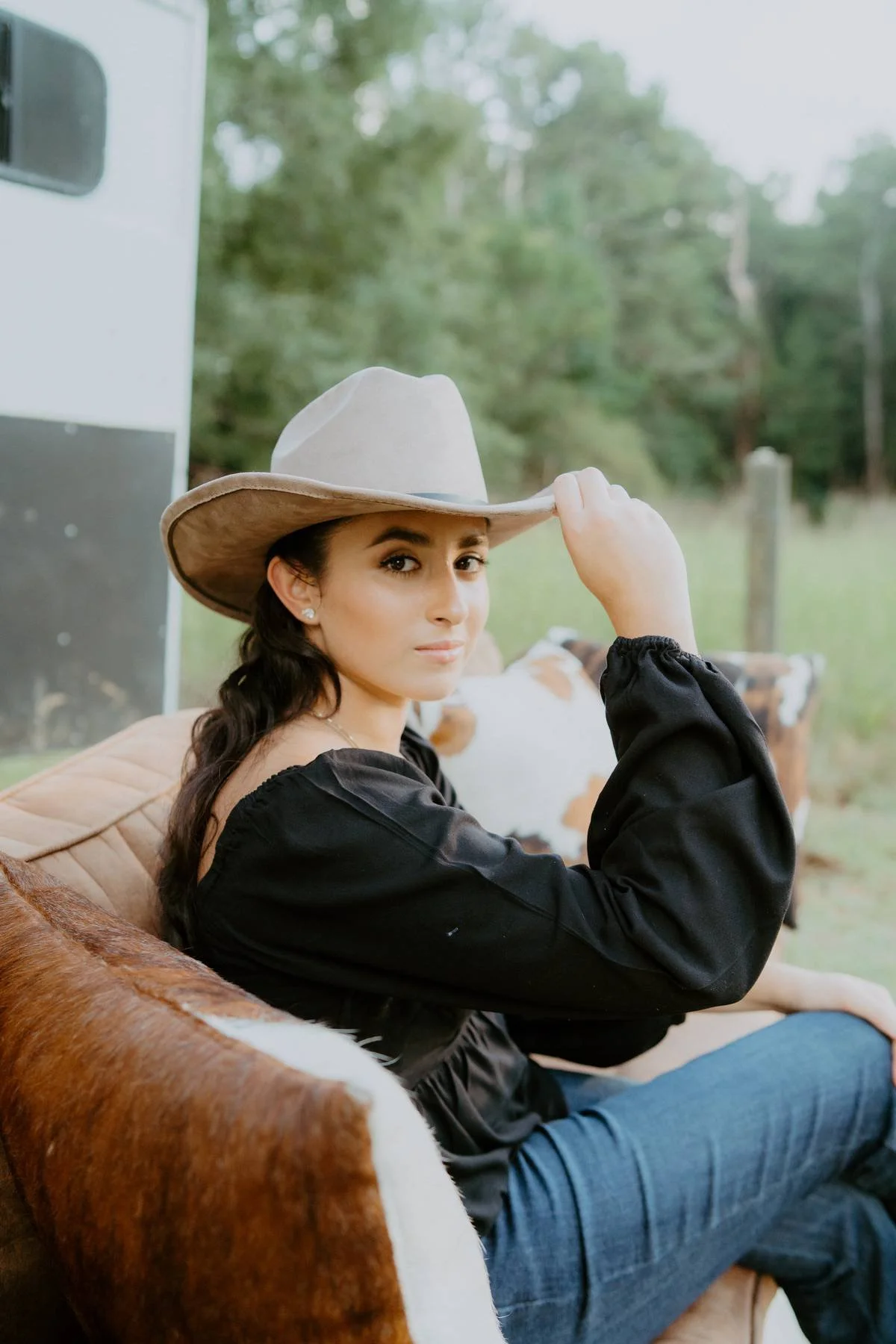 Woman sitting on a leather sofa outdoors, wearing a cowboy hat and a black long-sleeve shirt, with a cowhide pillow behind her, and greenery in the background.