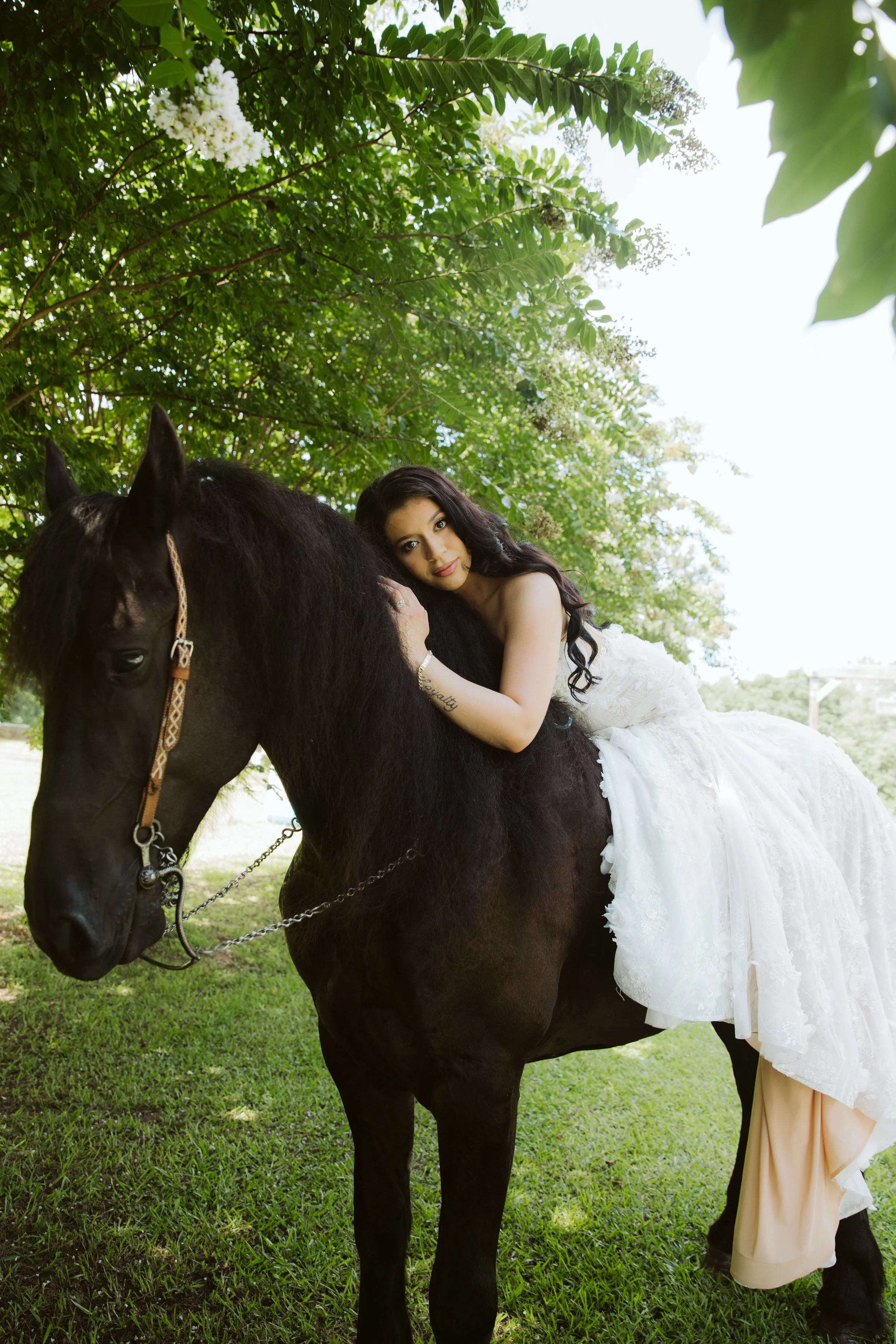 A woman in a white wedding dress lying on a black horse outdoors under green trees.