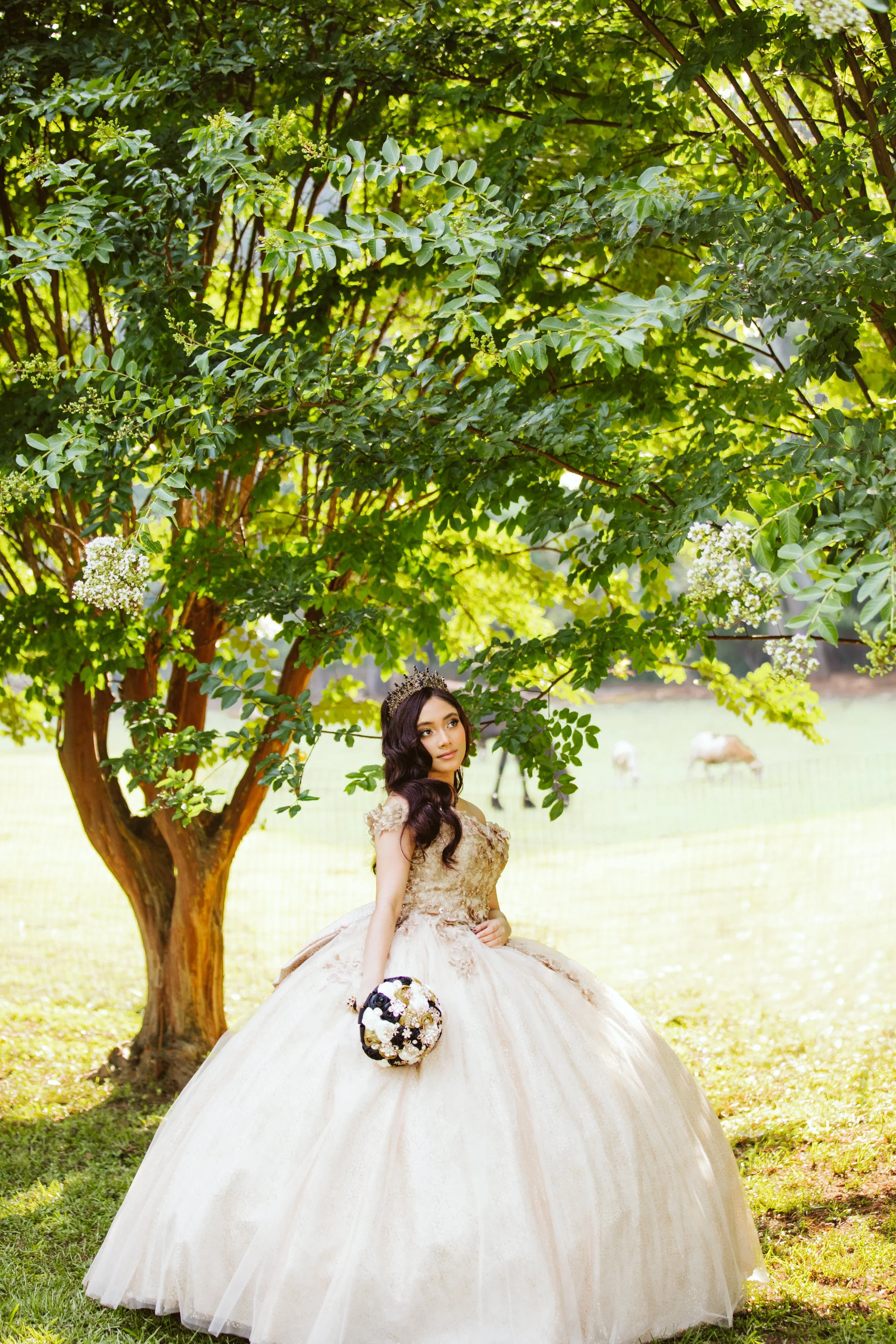 A woman in a ball gown sitting outdoors under a large leafy tree holding a bouquet of flowers.