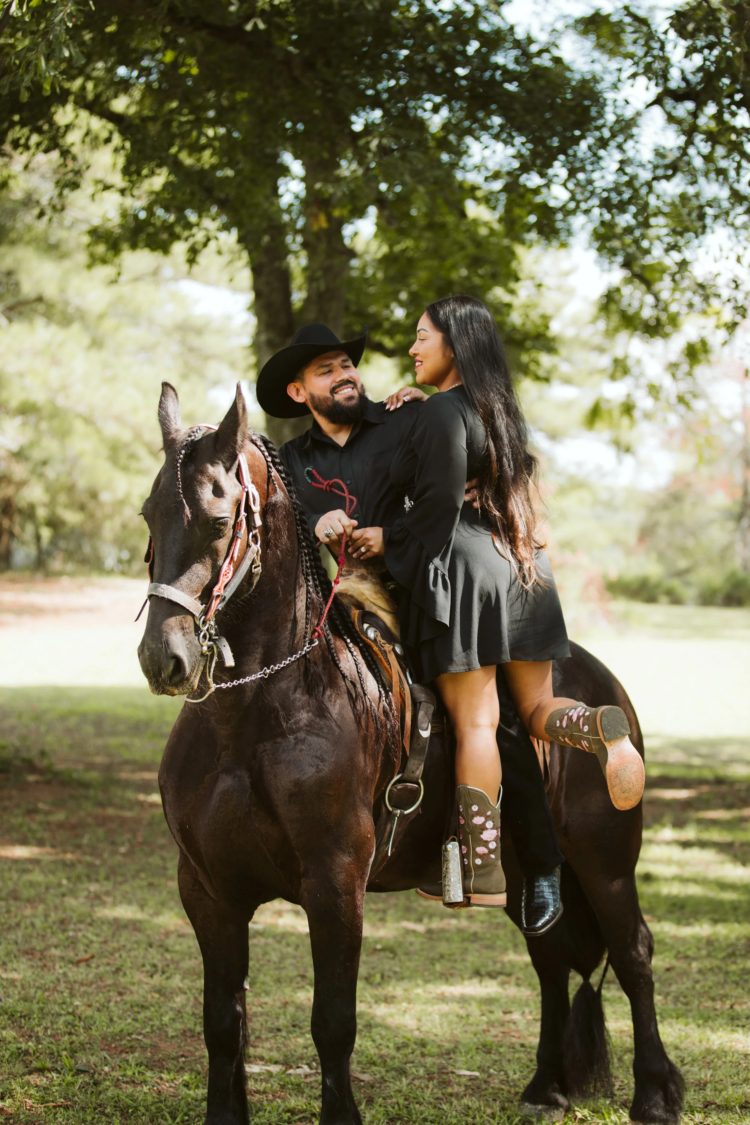 A man and woman riding a horse outdoors in a park with trees in the background. The man is wearing a cowboy hat and smiling at the woman, who is looking at him with a smile. The woman is wearing a black dress and sitting on the horse's back. The horse is dark brown with a black mane and tail, and has a saddle and bridle.