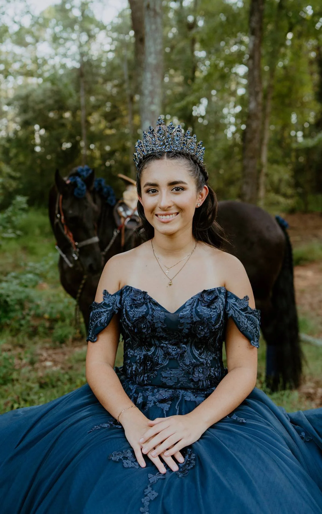 A young woman in a dark blue, off-shoulder gown sitting outdoors in a wooded area, wearing a matching tiara, with a black horse behind her, decorated with blue flowers in its mane.
