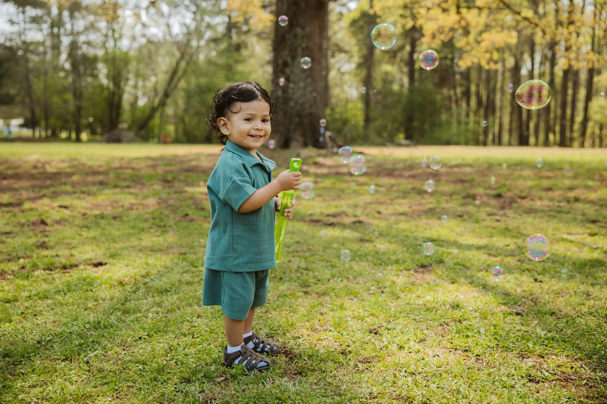 Young boy playing outdoors, holding a bubble wand, surrounded by floating soap bubbles, with a forest background.